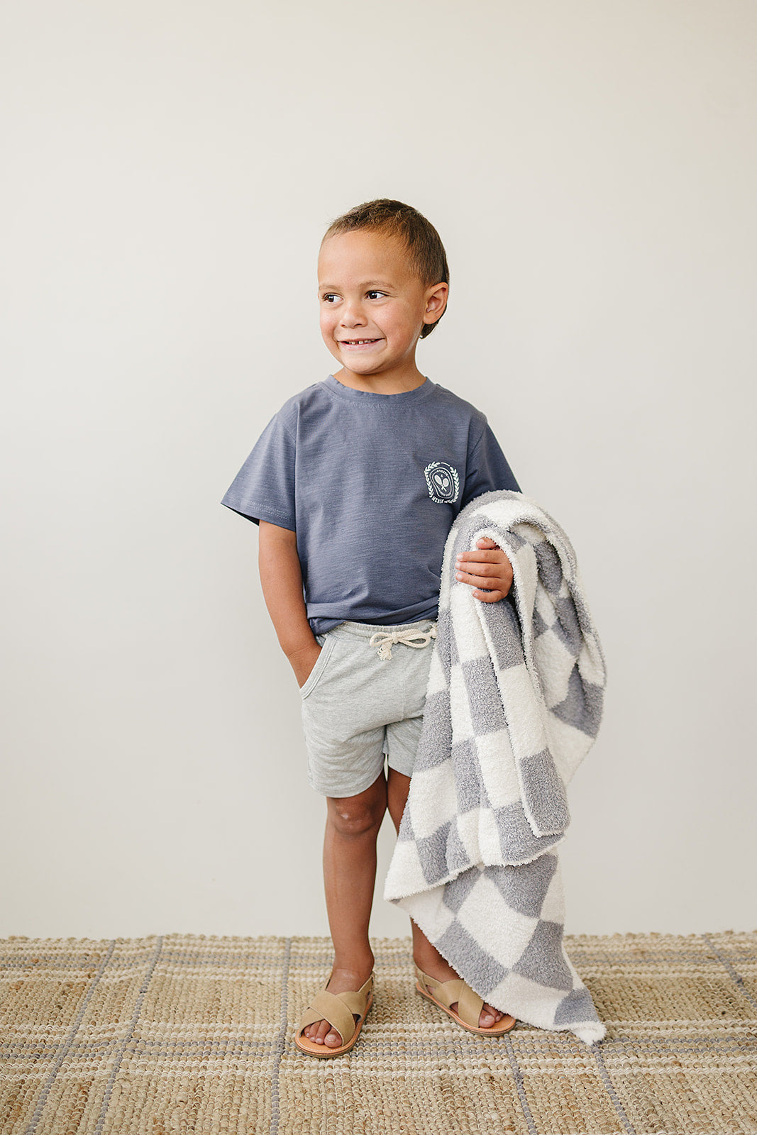Smiling child in a grey t-shirt and shorts holding a grey checkered plush blanket, showcasing modern style.