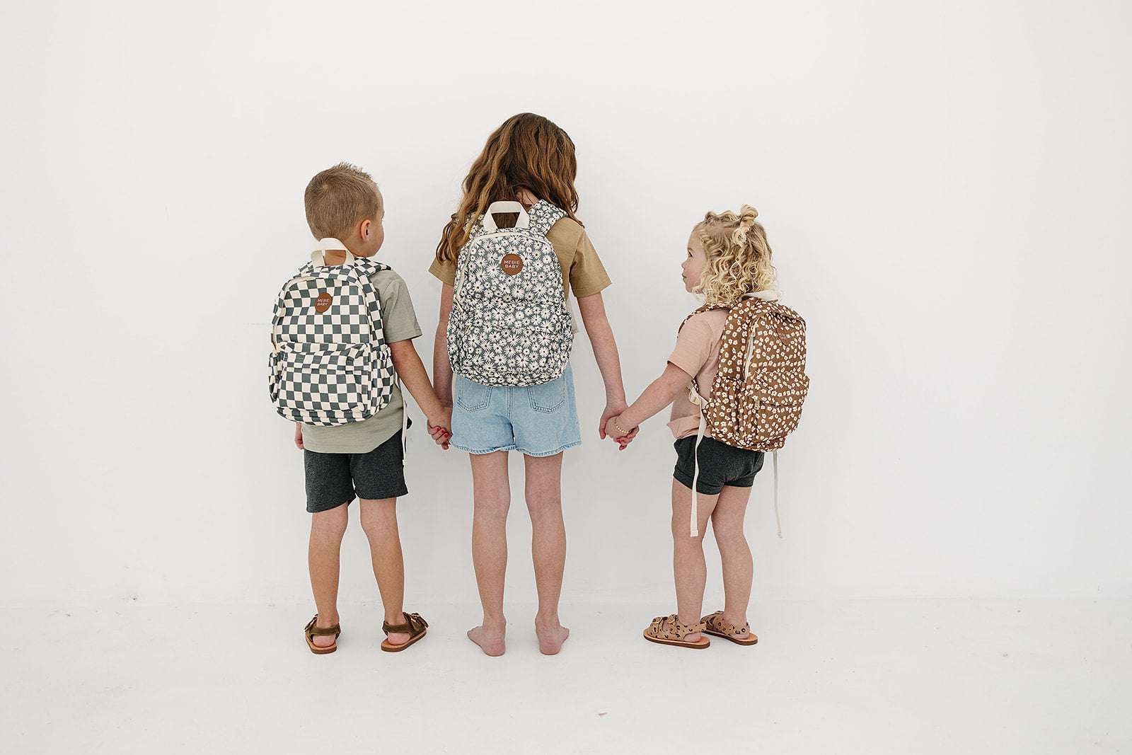 Three kids holding hands, each wearing stylish mini backpacks in playful patterns.