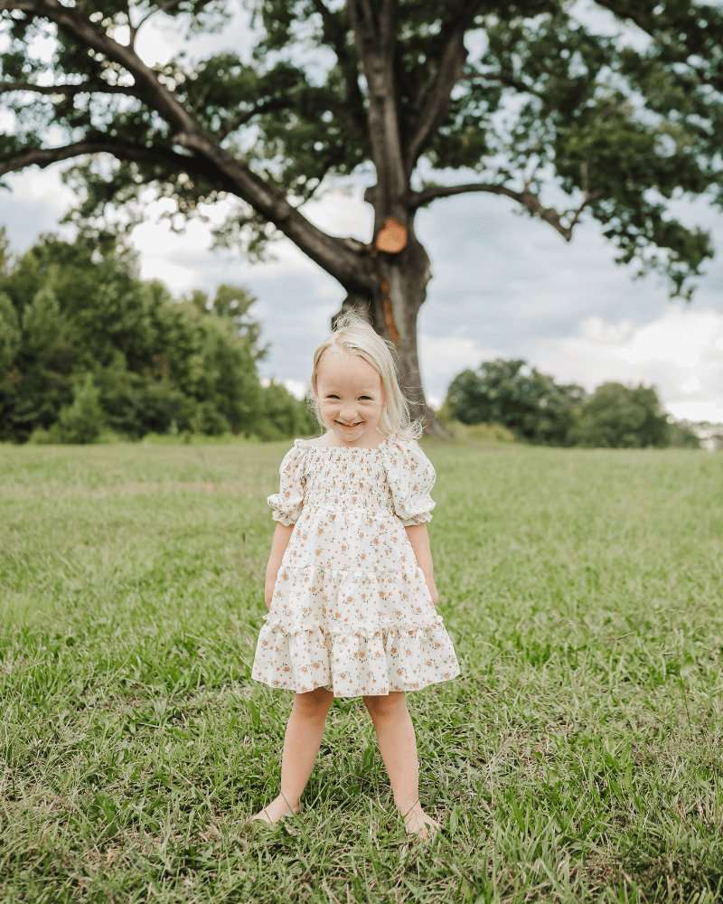 Smiling child in a yellow floral dress, enjoying a sunny day outdoors in a grassy field.