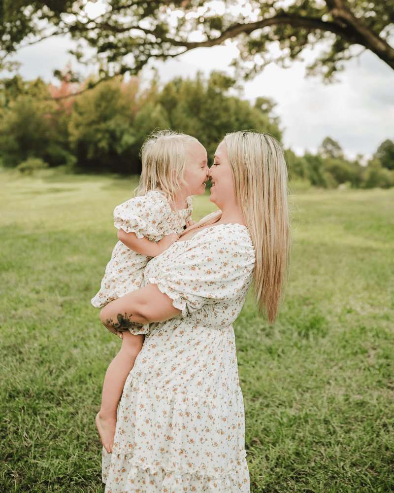 Mother and daughter smiling while wearing matching yellow floral dresses in a sunny outdoor setting.
