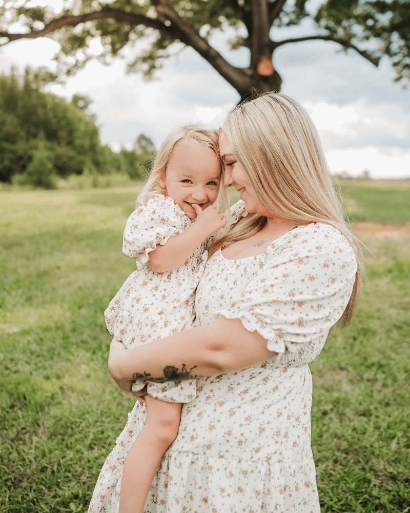 Mom and daughter in matching yellow floral dresses, enjoying a sunny day outdoors in a grassy field.