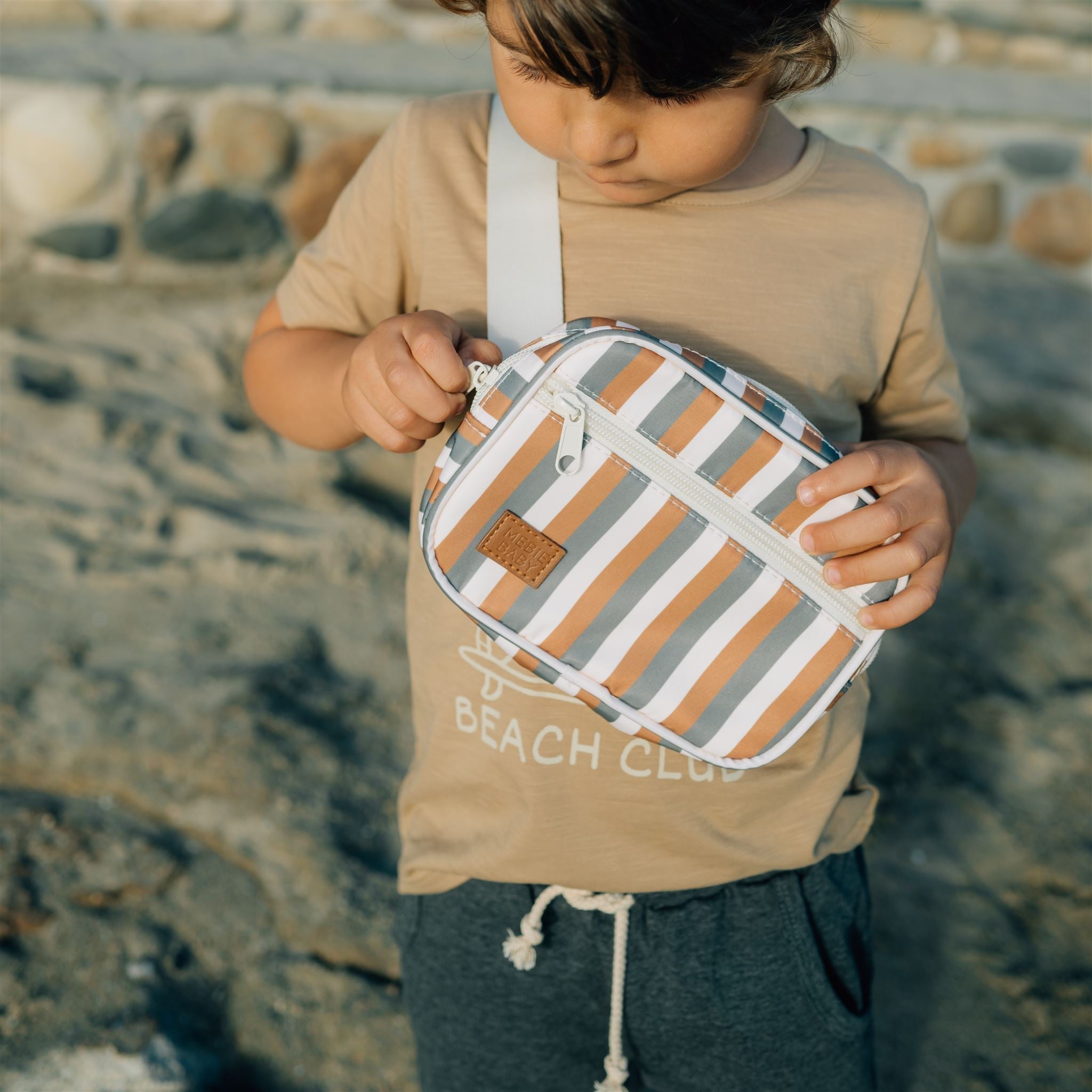 Child holding a Sunset Stripe Mini Fanny Pack with earthy tones and stripes on a beach background.