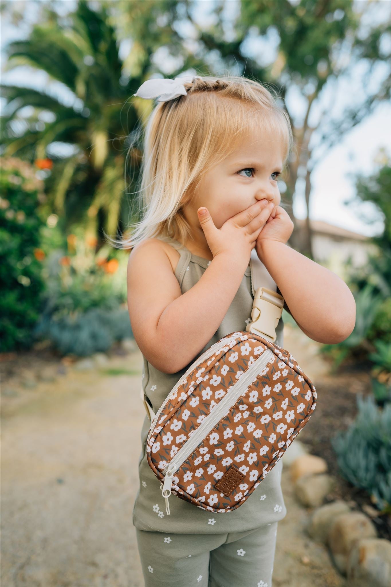 Toddler wearing a mustard floral mini fanny pack while playing outdoors in a garden setting.