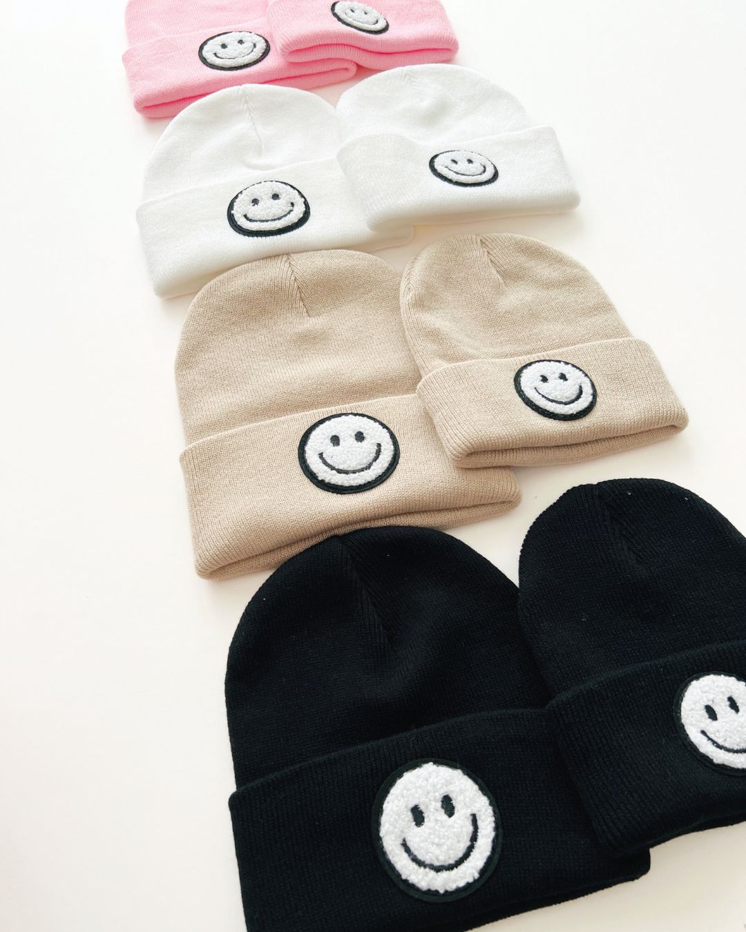 Collection of Smiley Beanies in pink, white, beige, and black with smiley face designs, perfect for cozy, stylish wear.