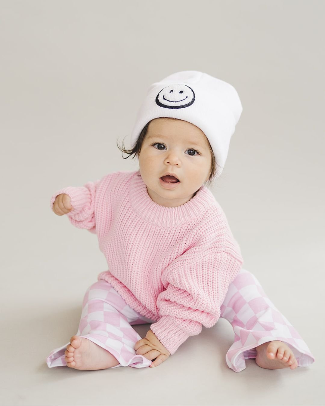 Toddler wearing a white Smiley Beanie and pink sweater, sitting on the floor for a cozy and stylish look.