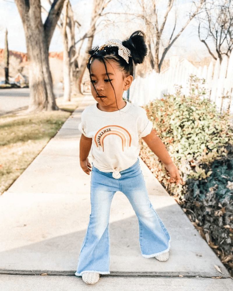 Toddler girl wearing light wash bell bottom jeans and a rainbow tee, enjoying a sunny day outdoors.