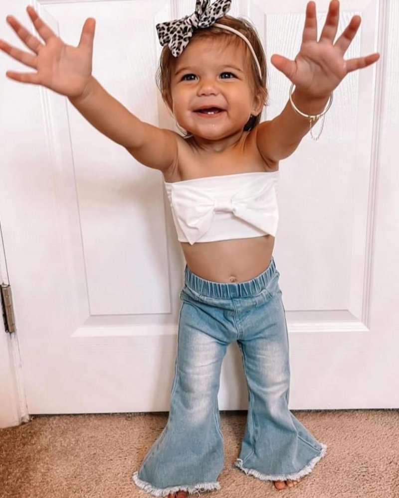 Toddler girl wearing light wash bell bottom jeans with a white top and a bow headband, showcasing a playful pose.