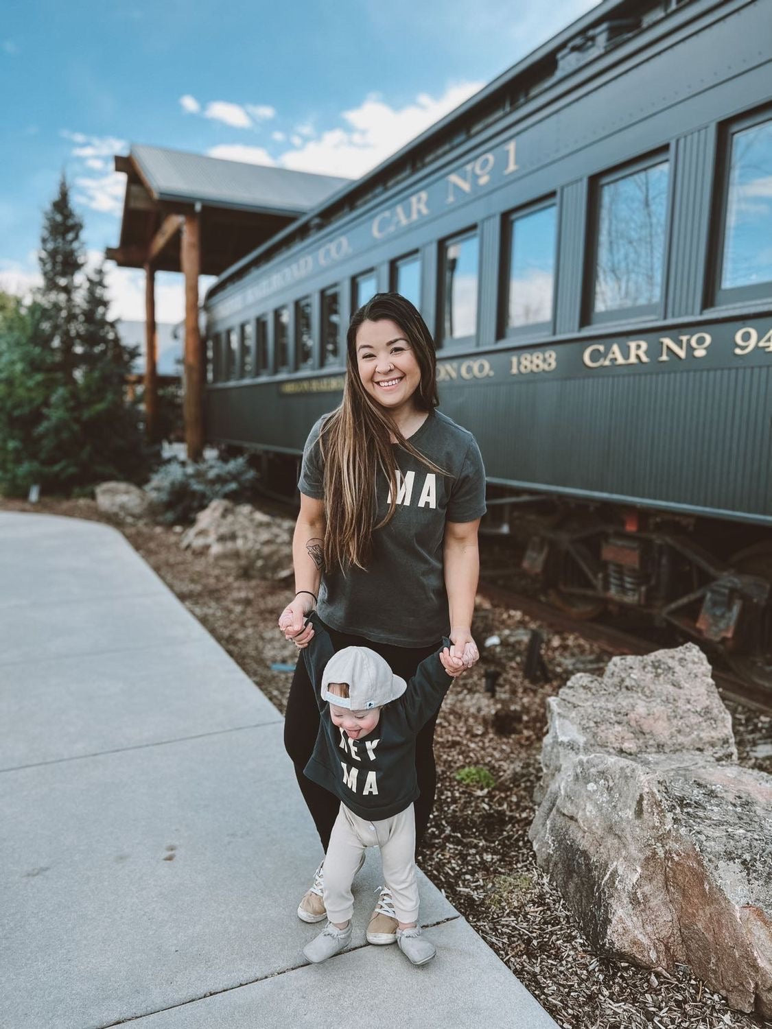 Mom and child wearing matching Hey Ma kids sweatshirt in black, enjoying a day out near a vintage train.