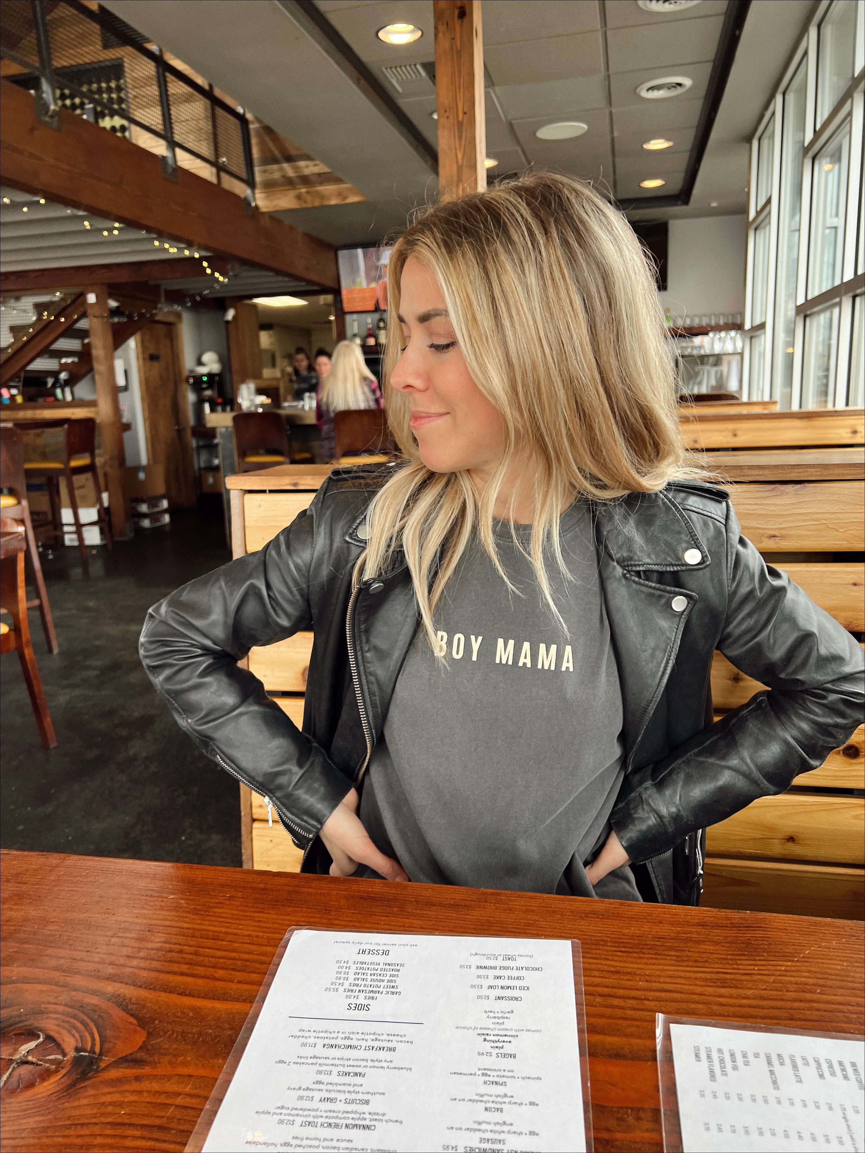 Woman wearing a gray 'Boy Mama' tee with a leather jacket in a cozy restaurant setting.