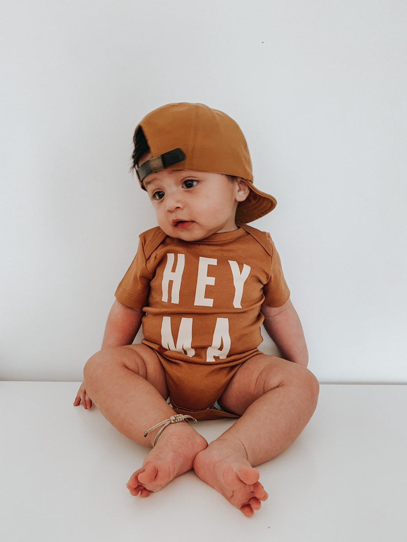 Adorable baby wearing a cream organic onesie with 'Hey Ma' slogan and a cap, perfect for trendy baby outfits.