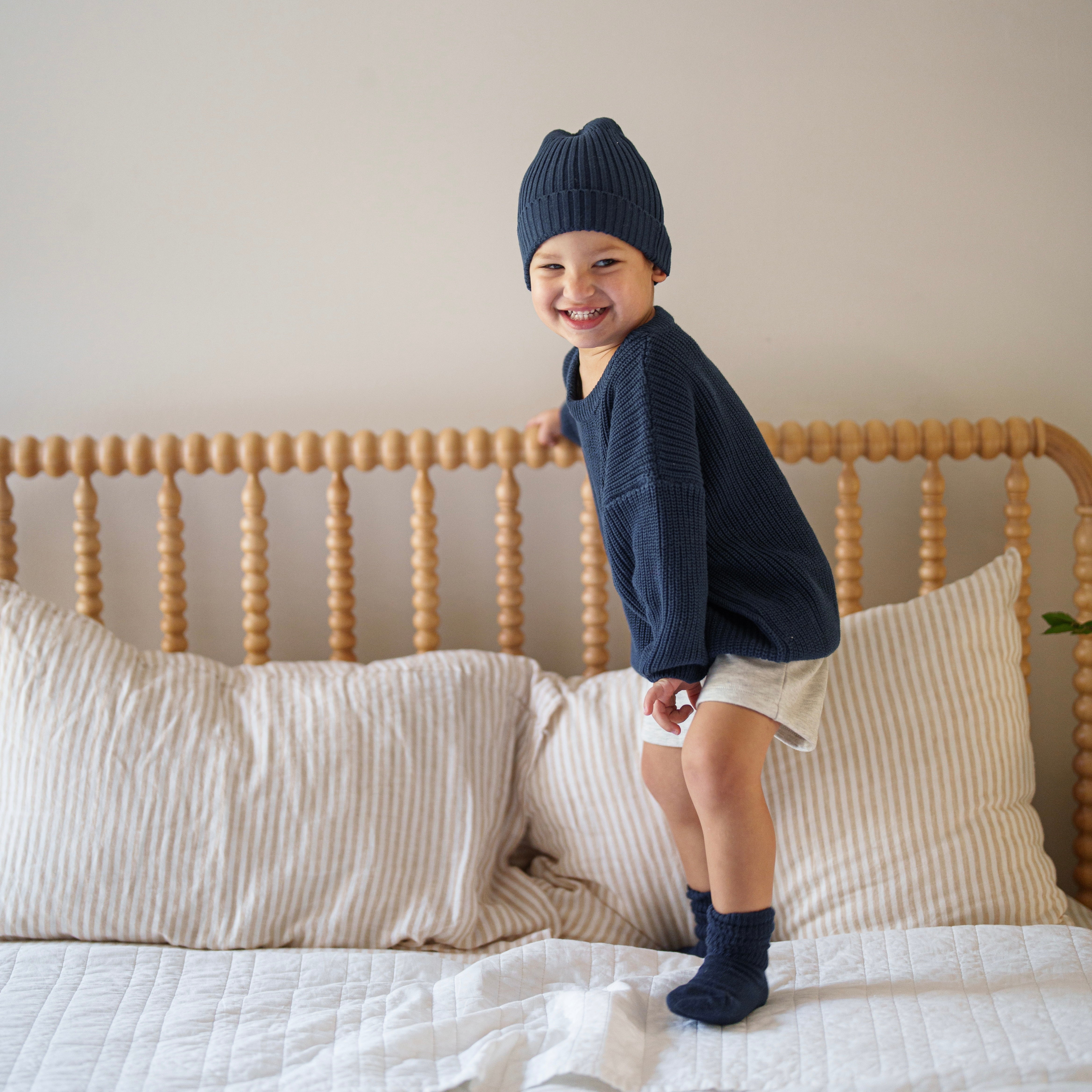 A happy child wearing an Organic Chunky Knit Sweater in navy, sitting on a bed with striped pillows.