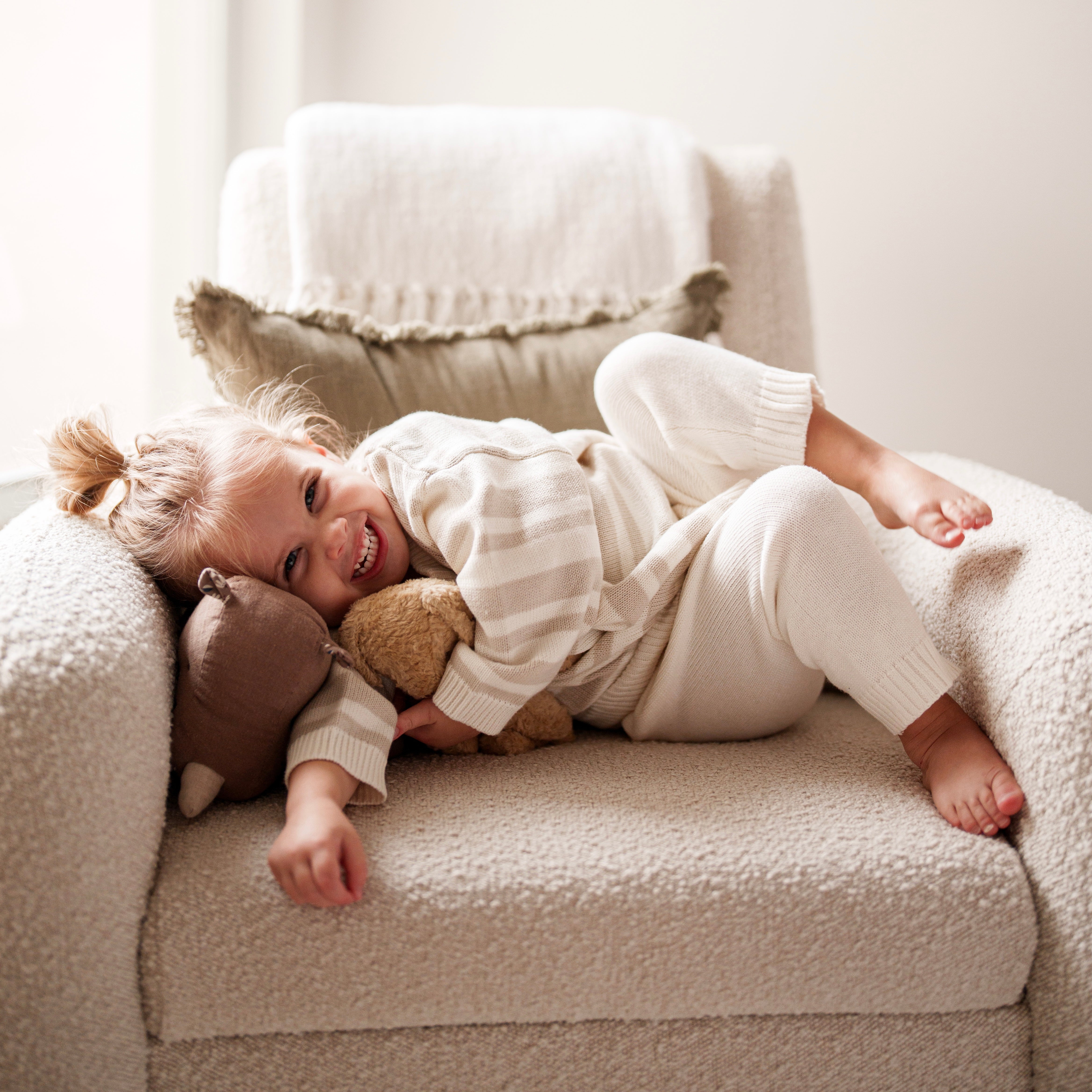Organic Knit Oversized Pant - Cotton Baby Bottoms worn by a smiling toddler on a cozy chair cuddling a stuffed animal.