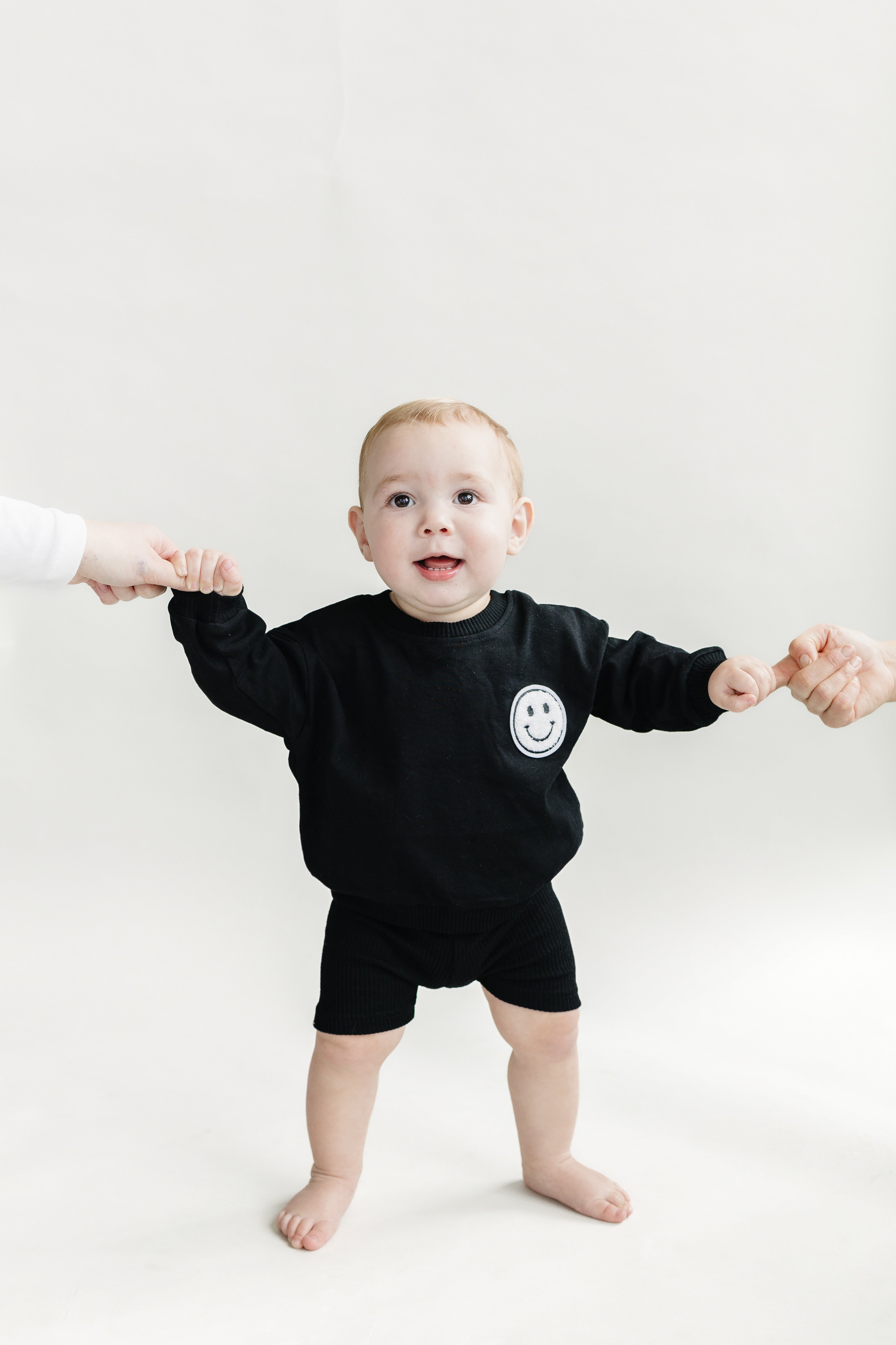 Baby wearing a black oversized Biker Shorts Set with a smiley face, standing and being held by two adults.