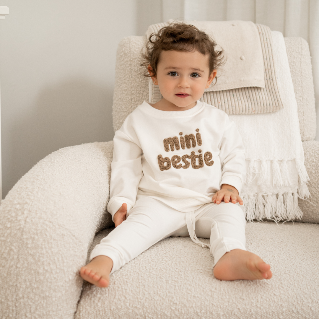 Toddler wearing a Mini Bestie Crewneck sweatshirt sitting on a cozy chair, showcasing its comfy design and fuzzy embroidery.