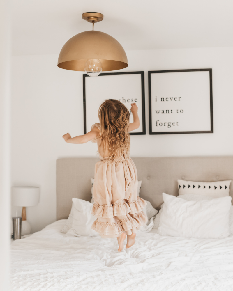 Girl in a champagne dress jumping on a bed, showcasing the playful elegance of the Nellie Ruffle Maxi Dress.