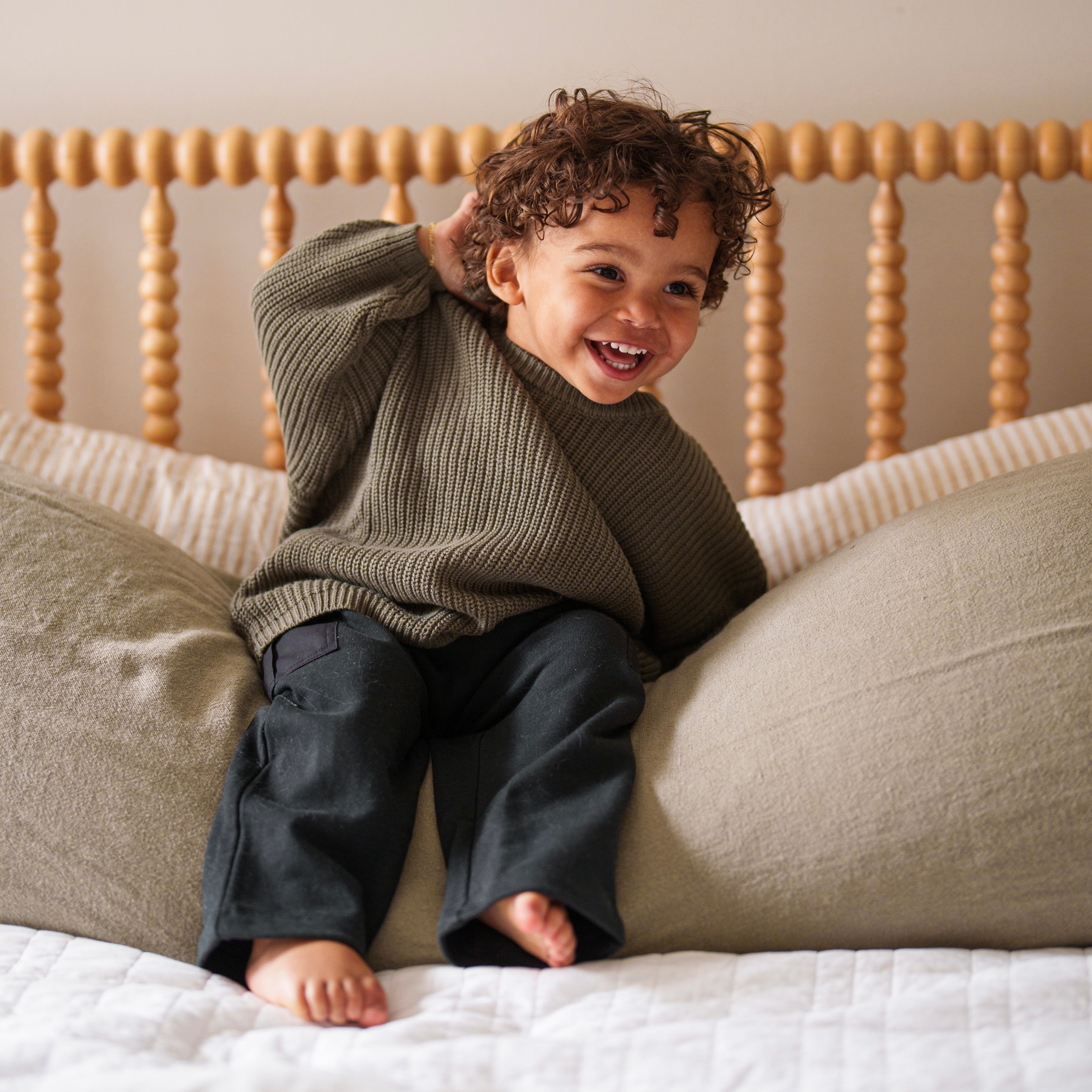 Happy child wearing Organic Chunky Knit Sweater and casual pants, sitting on bed with pillows.