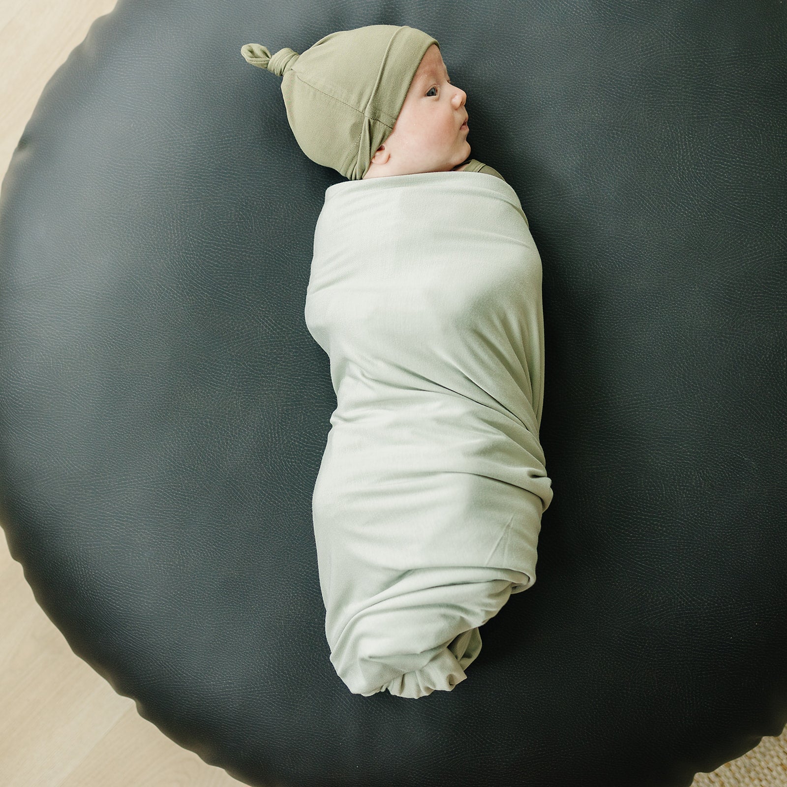 Baby wrapped in a Bamboo Stretch Swaddle Sage on a round black cushion, showcasing comfort and style.
