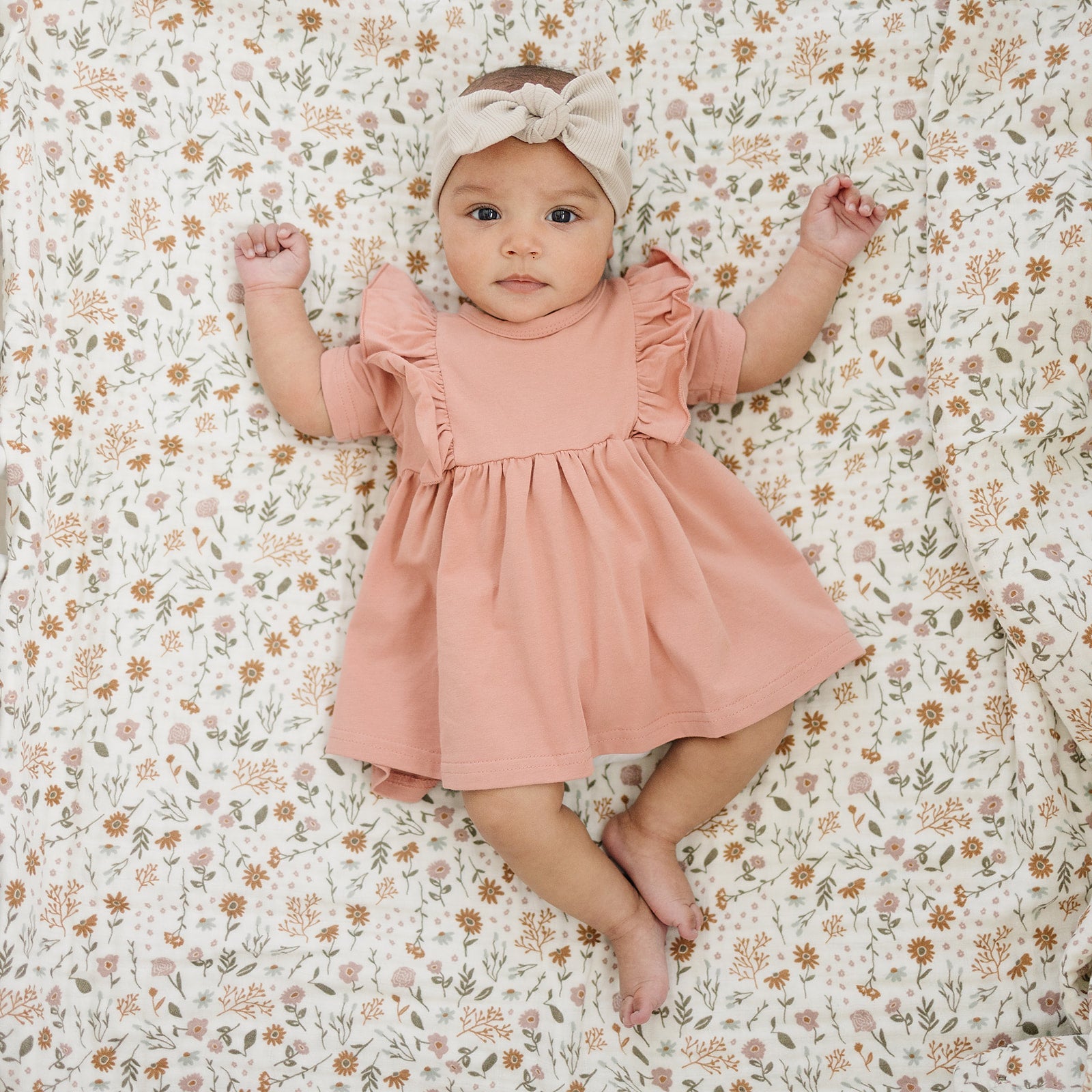 Baby girl in a pink dress with ruffled sleeves and a beige head wrap on a floral blanket.
