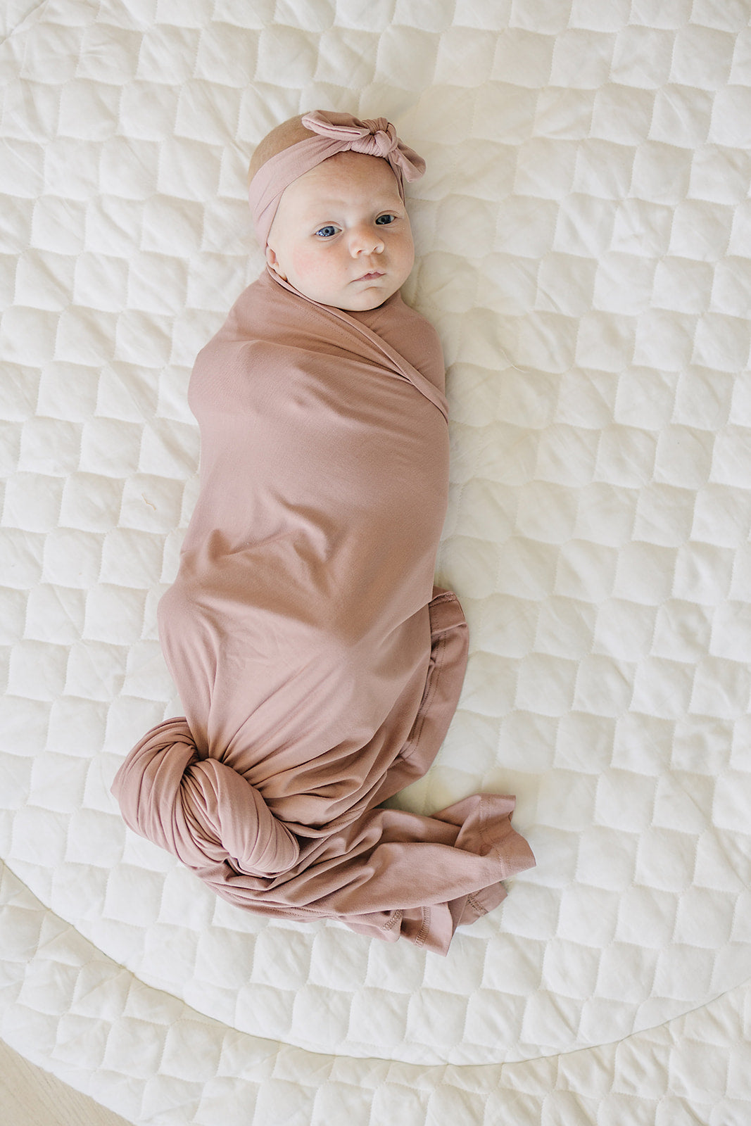 Bamboo Stretch Swaddle Dusty Rose wrapped around a baby on a quilted blanket, showcasing its softness and style.