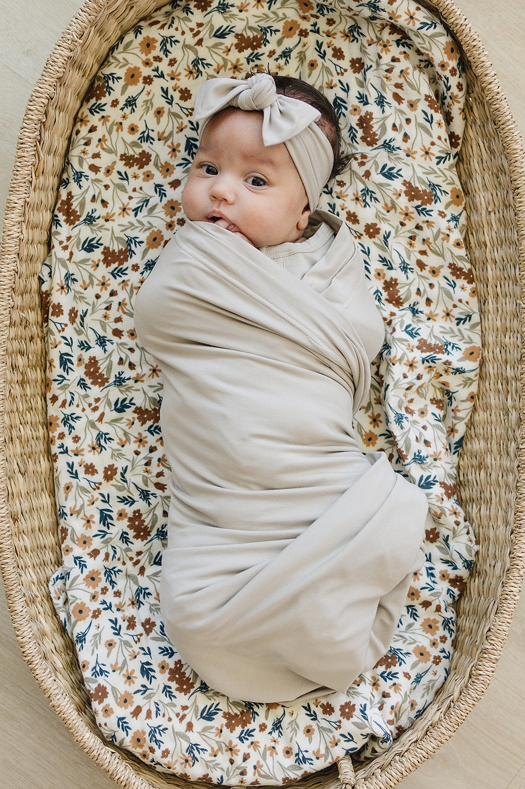 Bamboo Stretch Swaddle Oatmeal wrapped baby in a floral bassinet, showcasing the soft and neutral design.