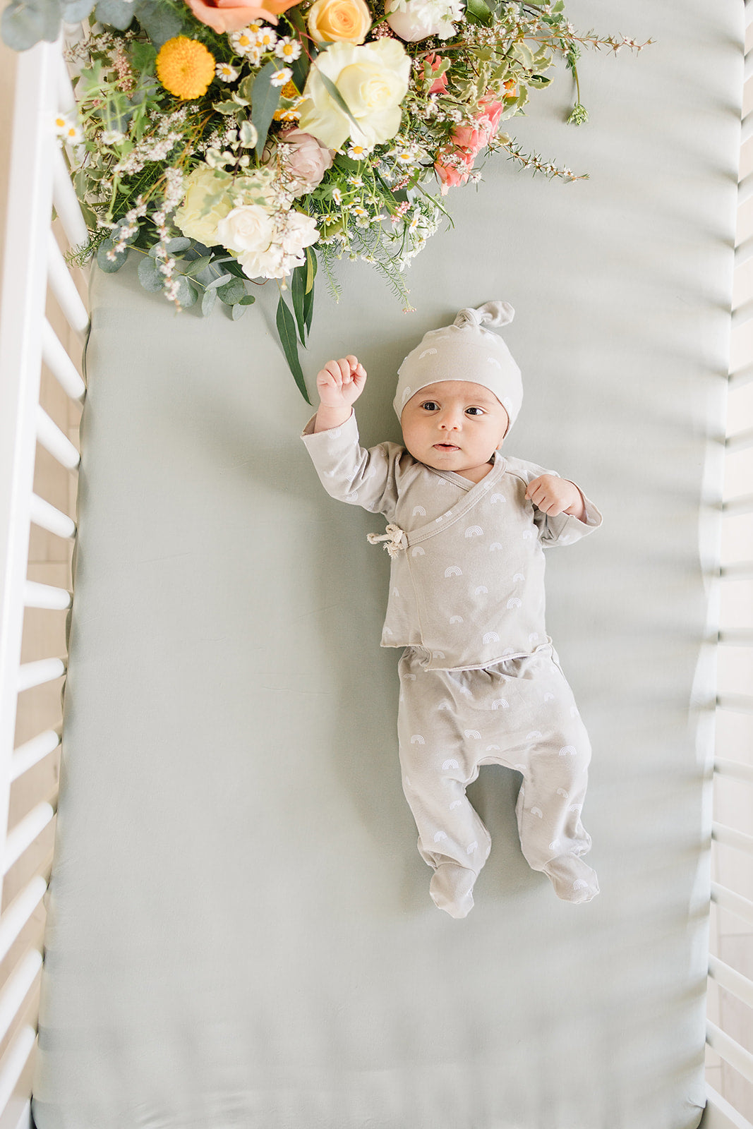 Baby lying on a bamboo crib sheet sage in a crib, surrounded by a floral arrangement.