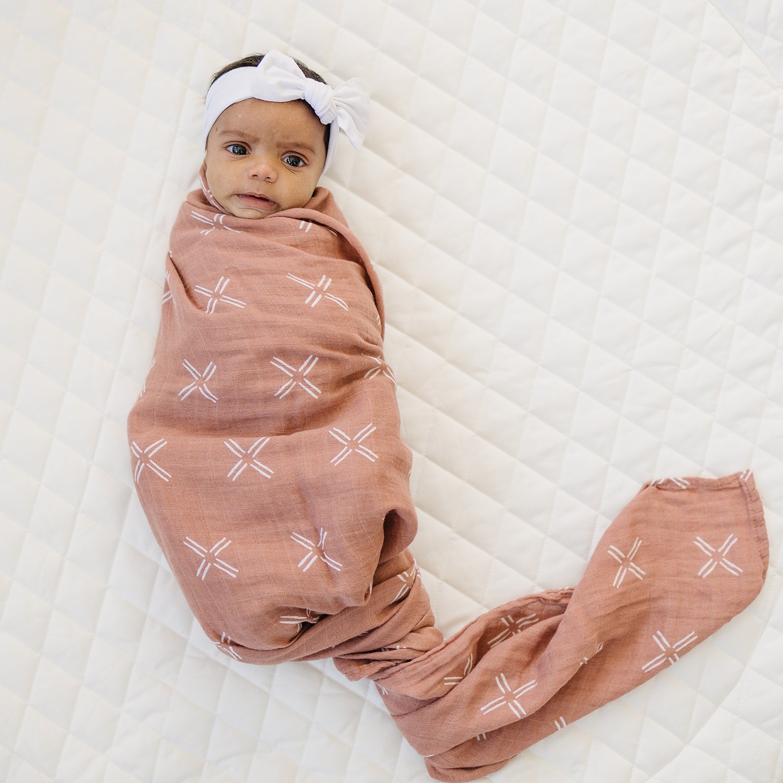 Baby wrapped in Just Peachy Muslin Swaddle Blanket on quilted surface, showcasing breathable cotton fabric.