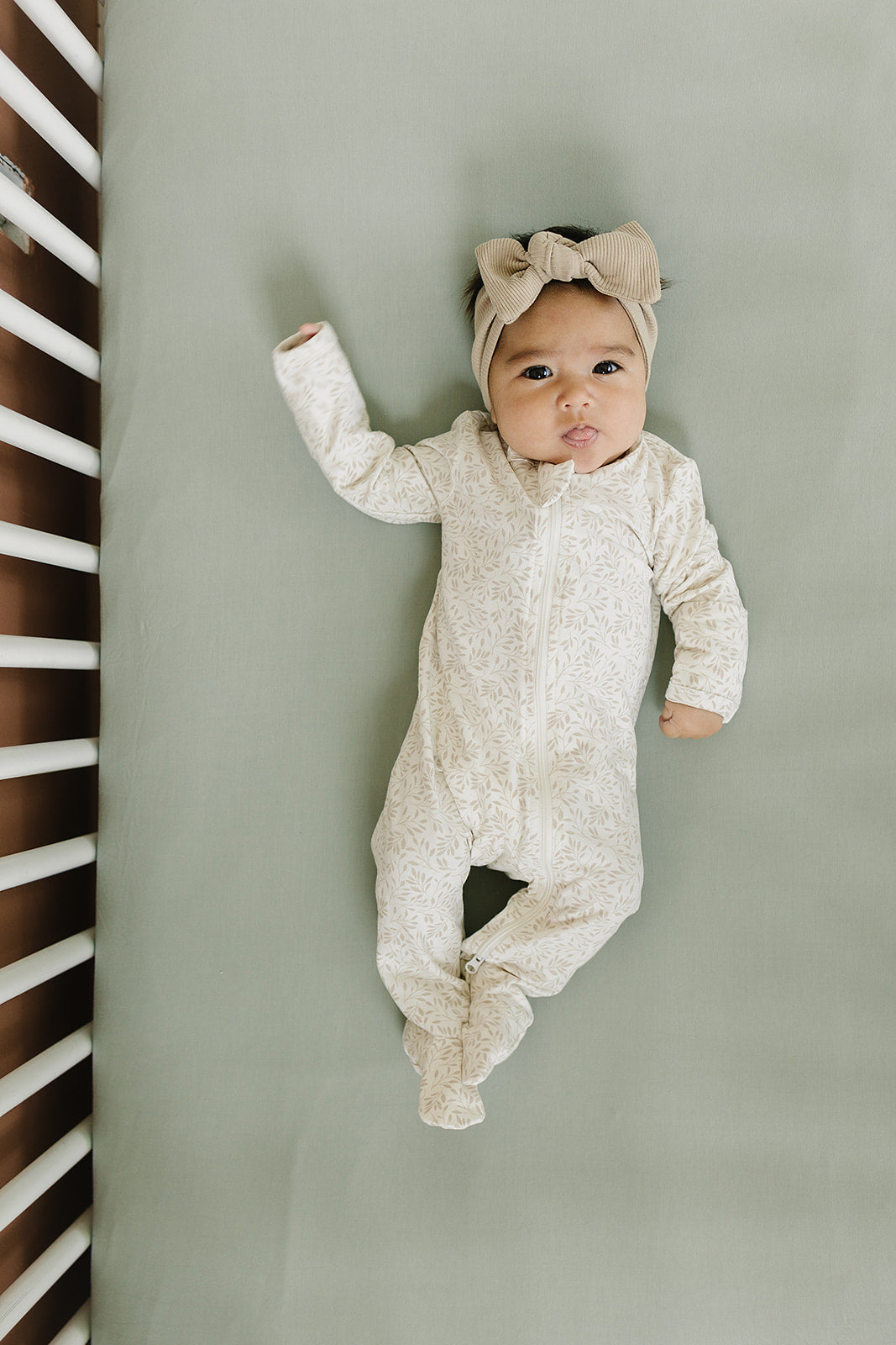 Baby resting on a bamboo crib sheet in sage color, wearing a cute onesie and headband.
