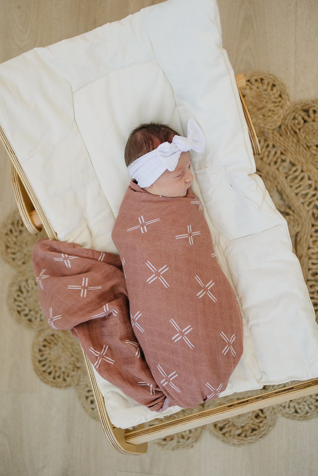Baby swaddled in a stylish muslin blanket on a bassinet with a headband, showcasing soft and cozy design.