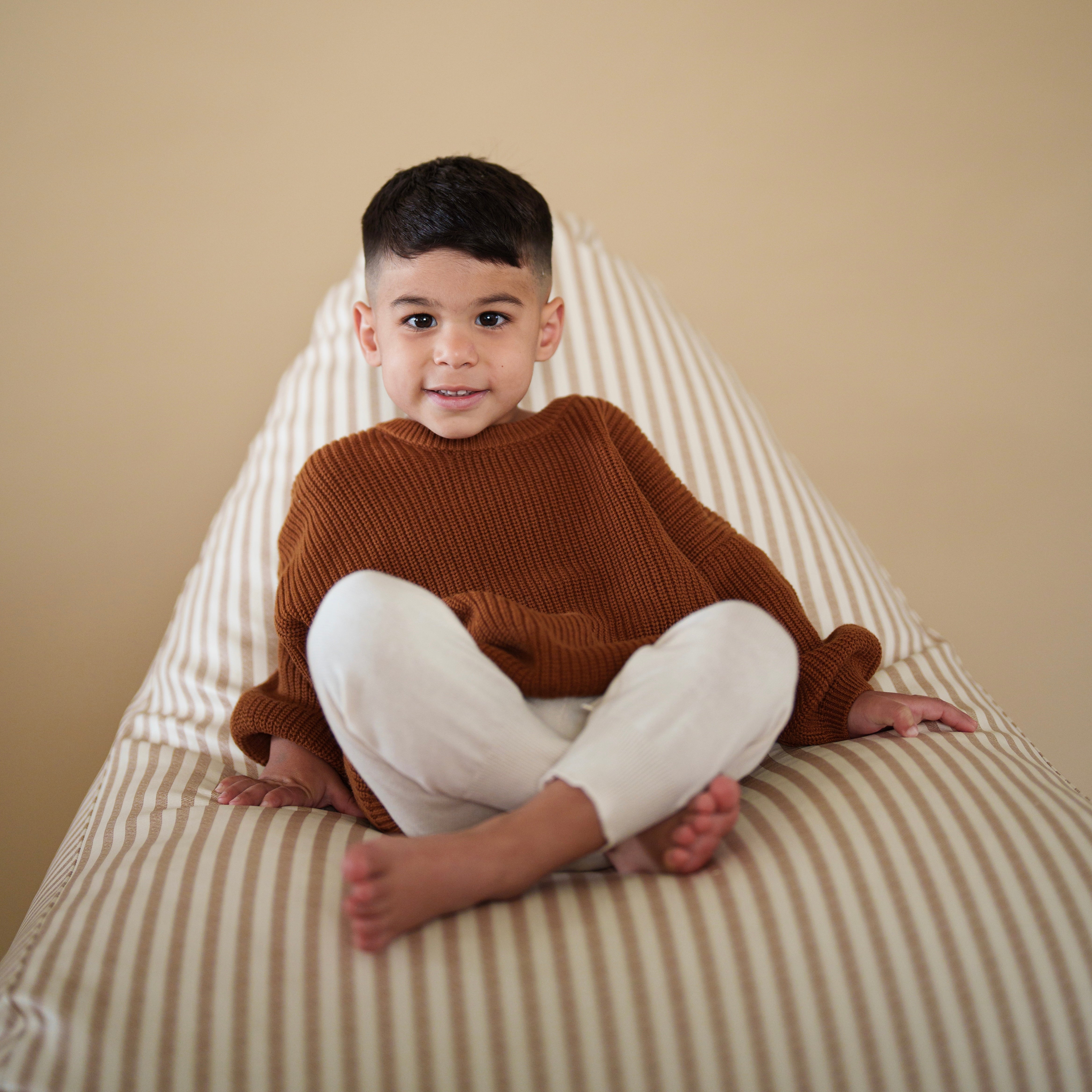 Child wearing Organic Chunky Knit Sweater in Coffee Bean color, sitting comfortably on a striped surface.