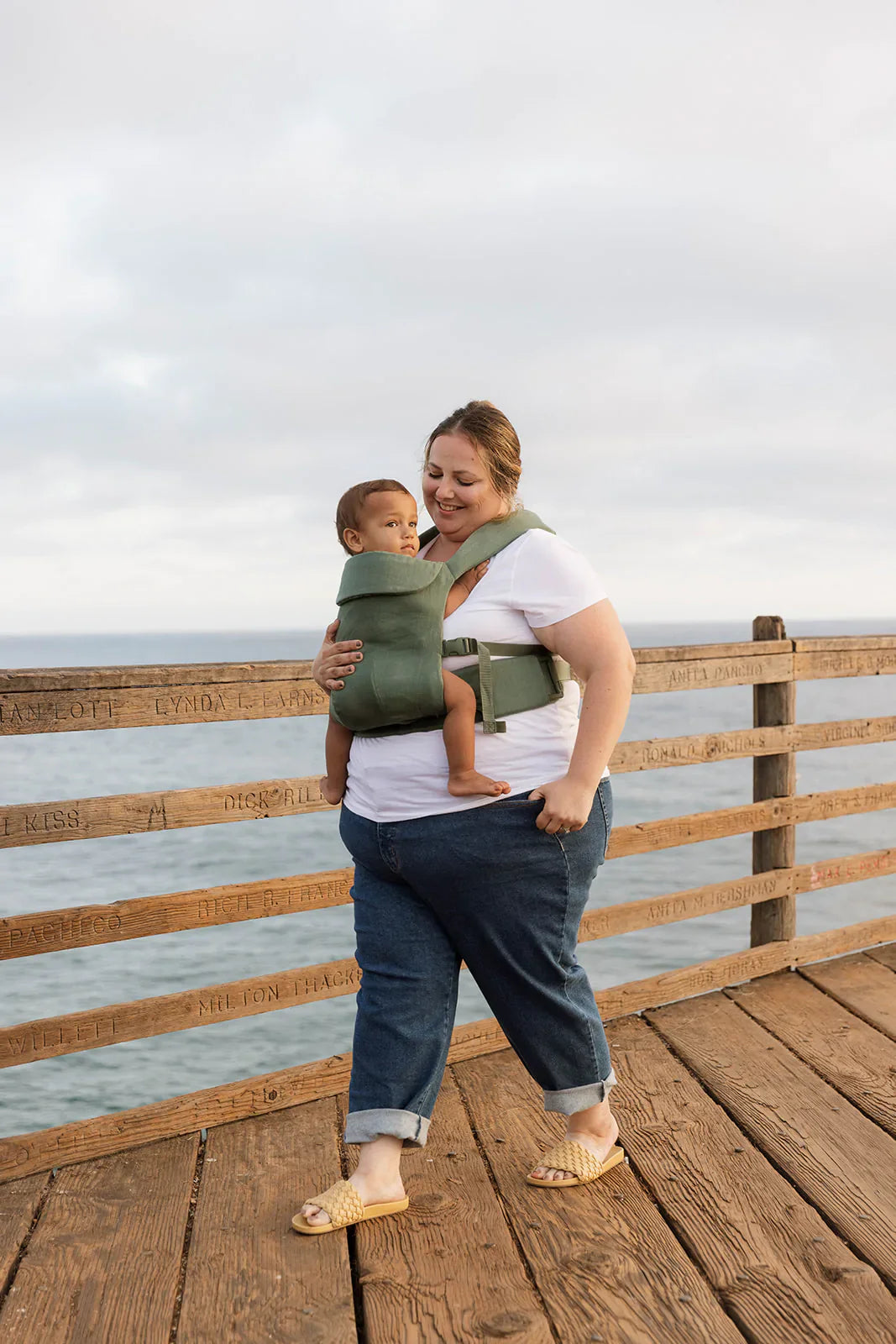 Woman carrying a child in dusky sage green Acadian aerial carrier on a seaside wooden pier.
