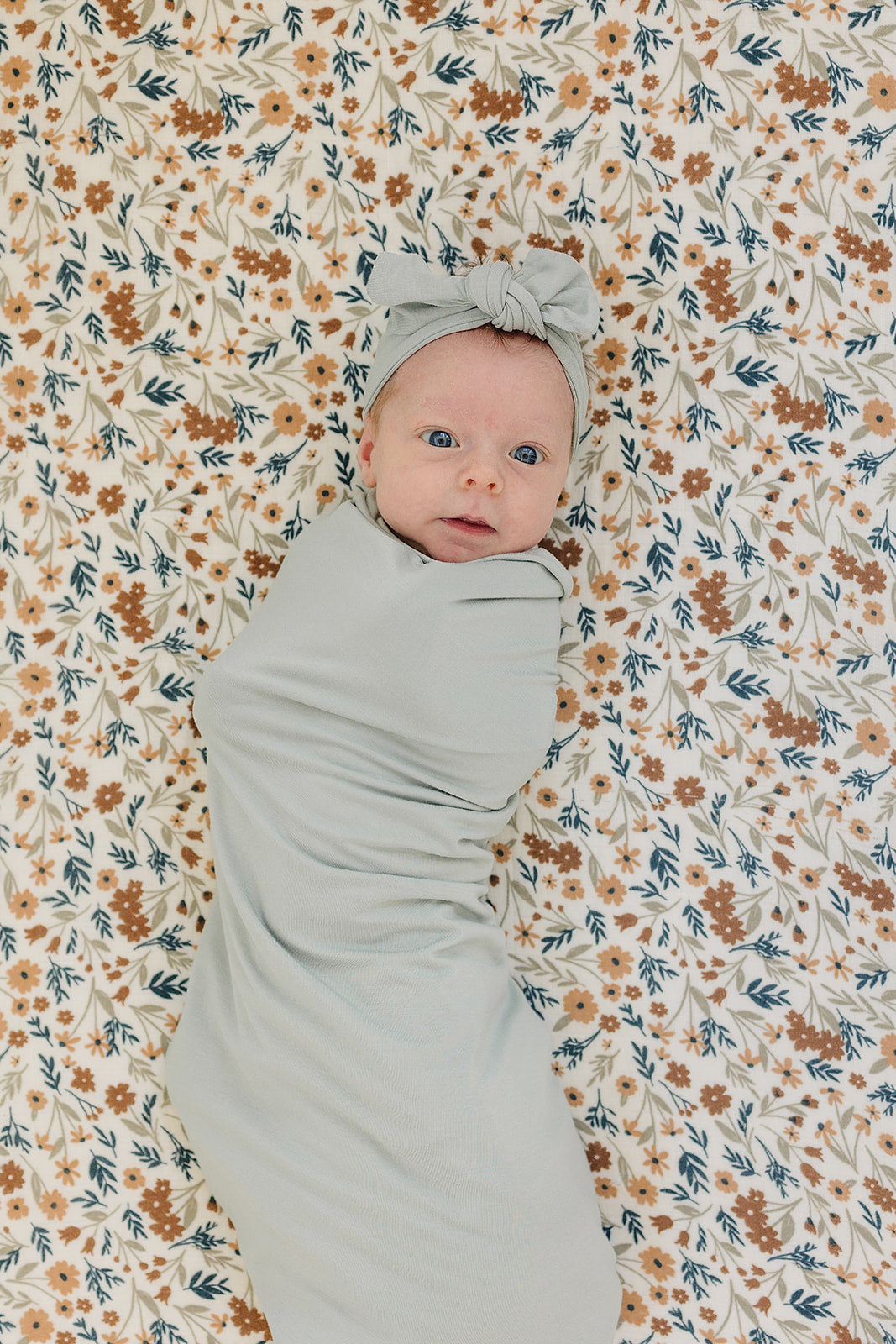 Bamboo Stretch Swaddle Sage wrapped around a baby on floral background, showcasing soft and breathable fabric.