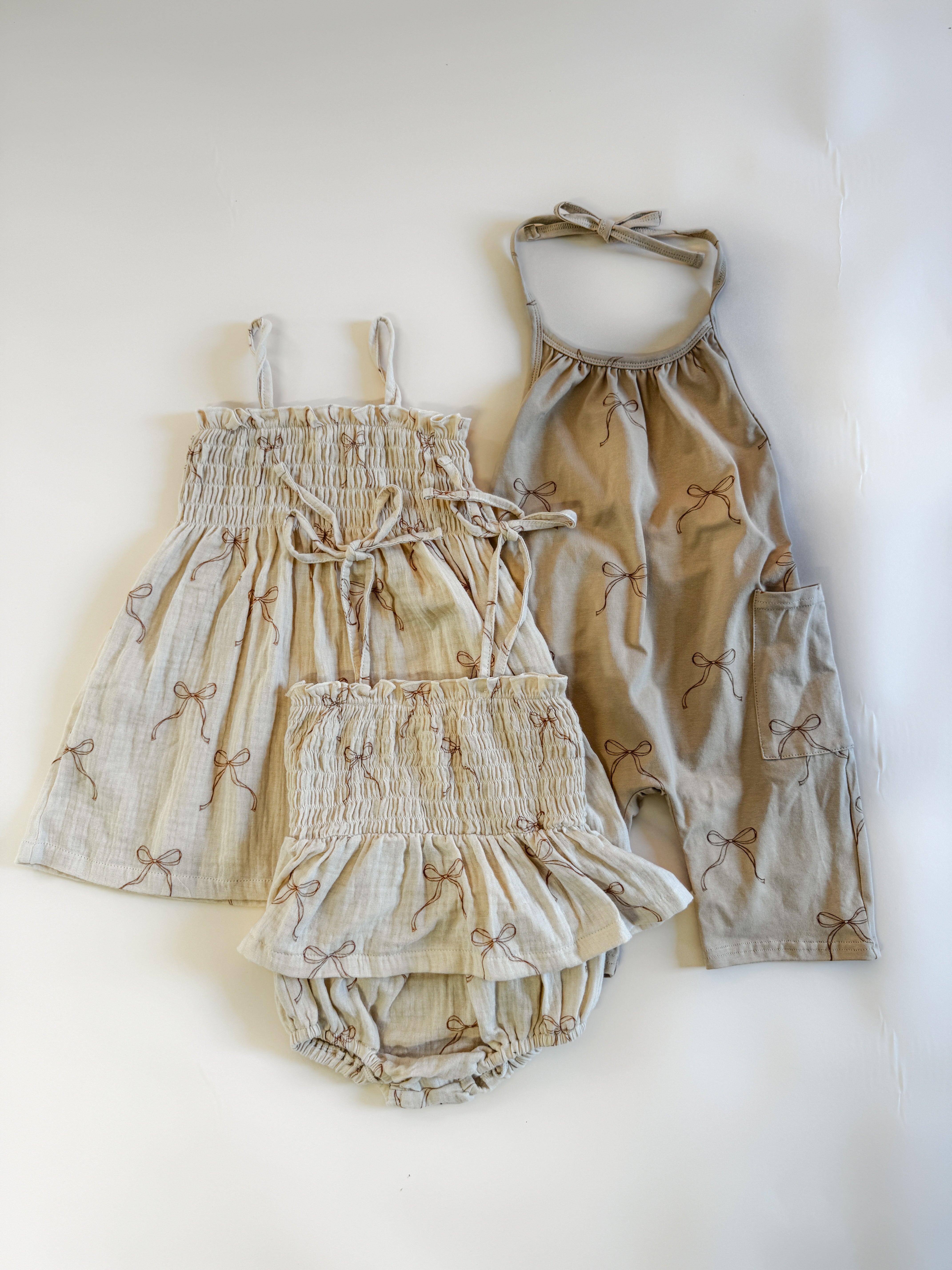 Set of three children's outfits featuring beige fabric with bow prints, including a smocked dress, romper, and bloomers.