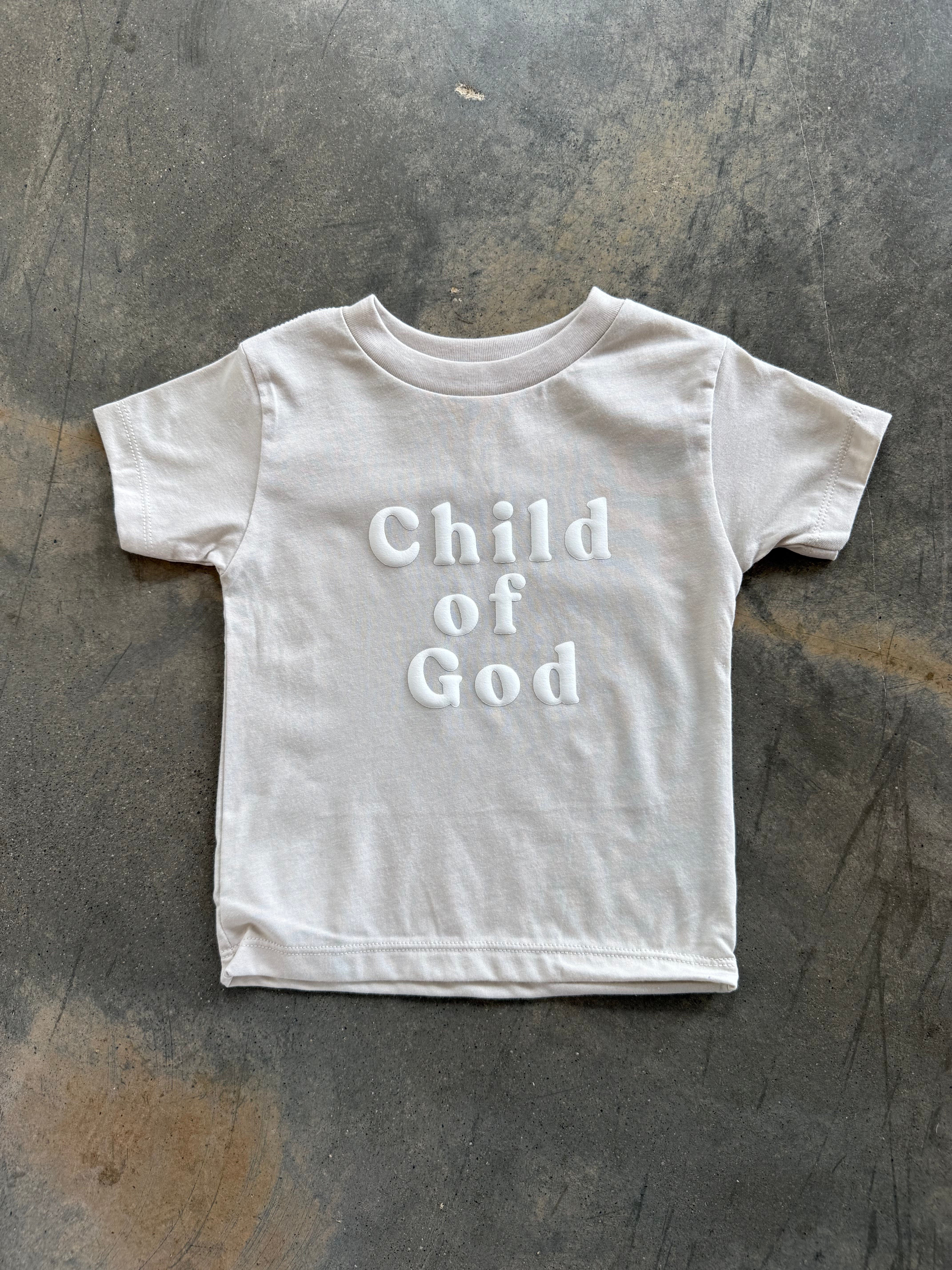 Child of God Kids Tee White with 'Child of God' text on a textured gray surface