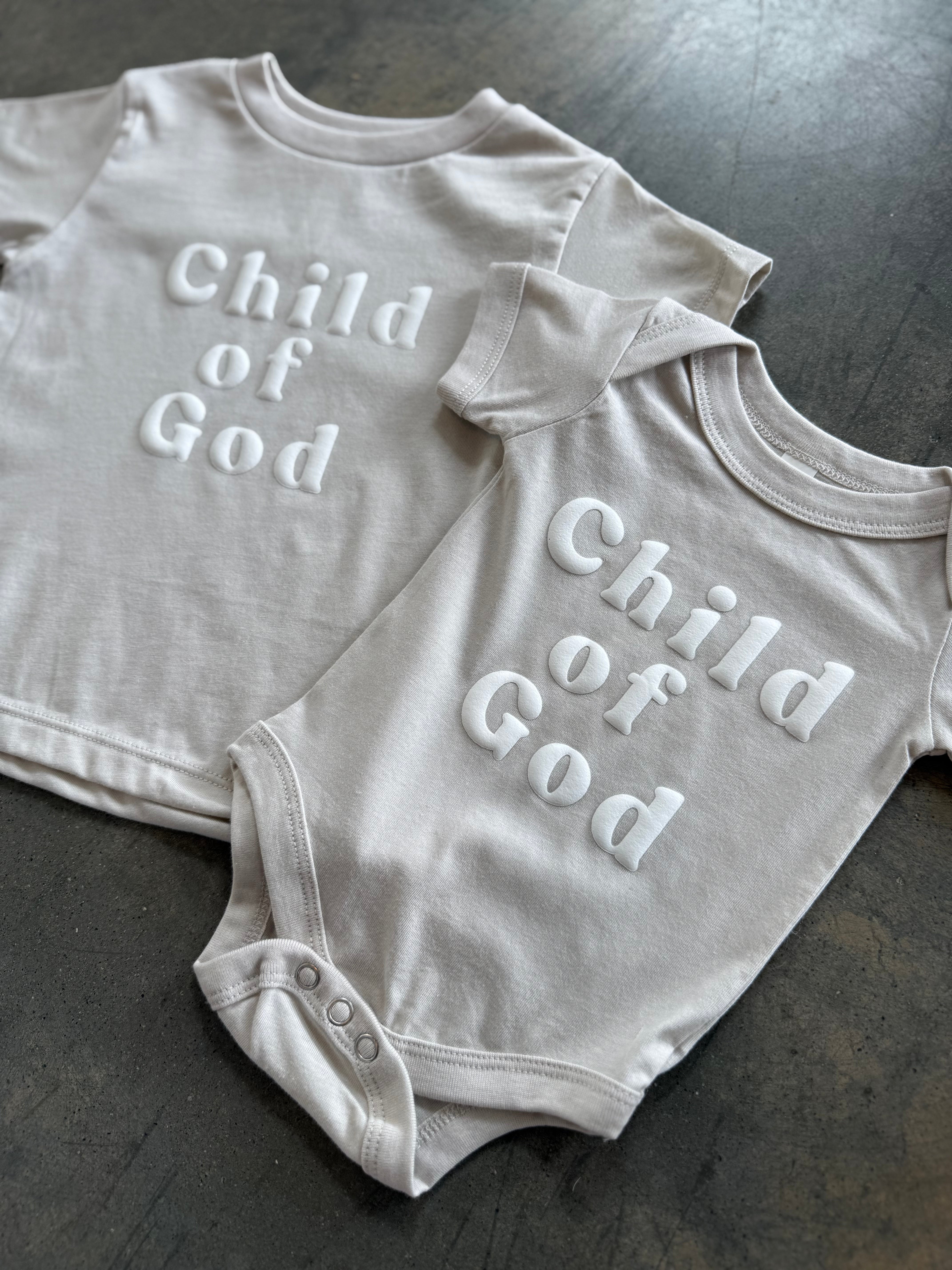 Child of God Kids Tee with Onesie on grey concrete floor at an angle