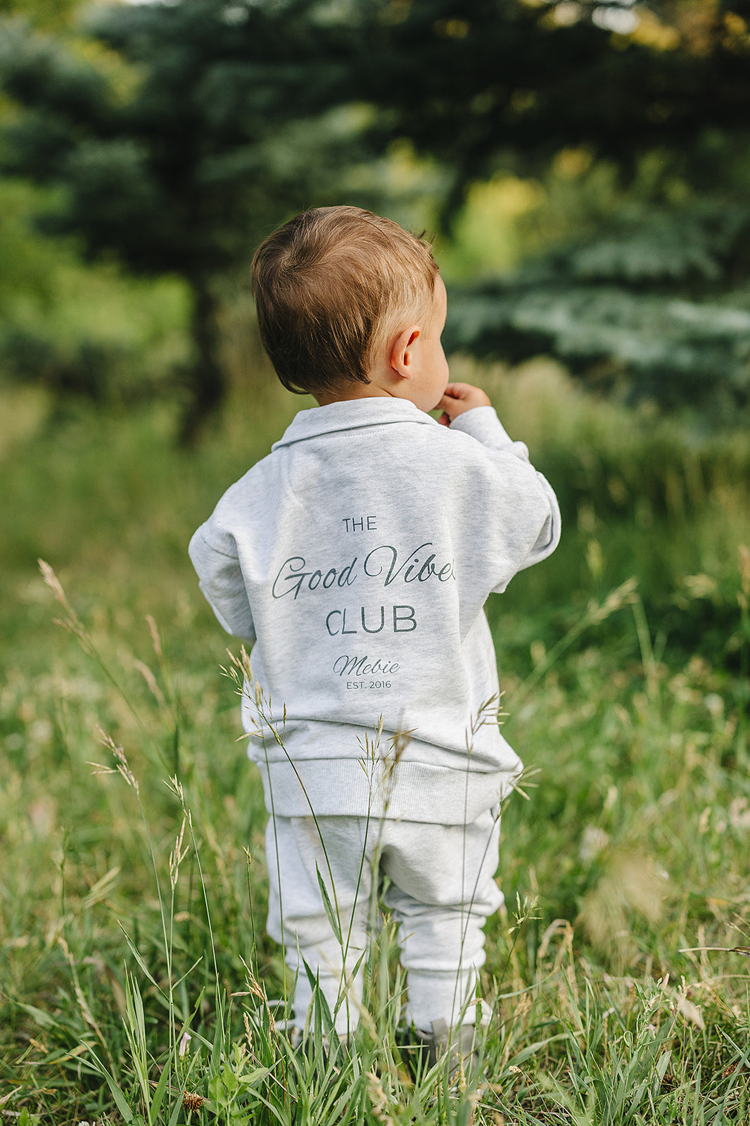 Toddler wearing unisex French terry quarter zip outfit in grey, featuring 'Good Vibes Club' design, surrounded by greenery.