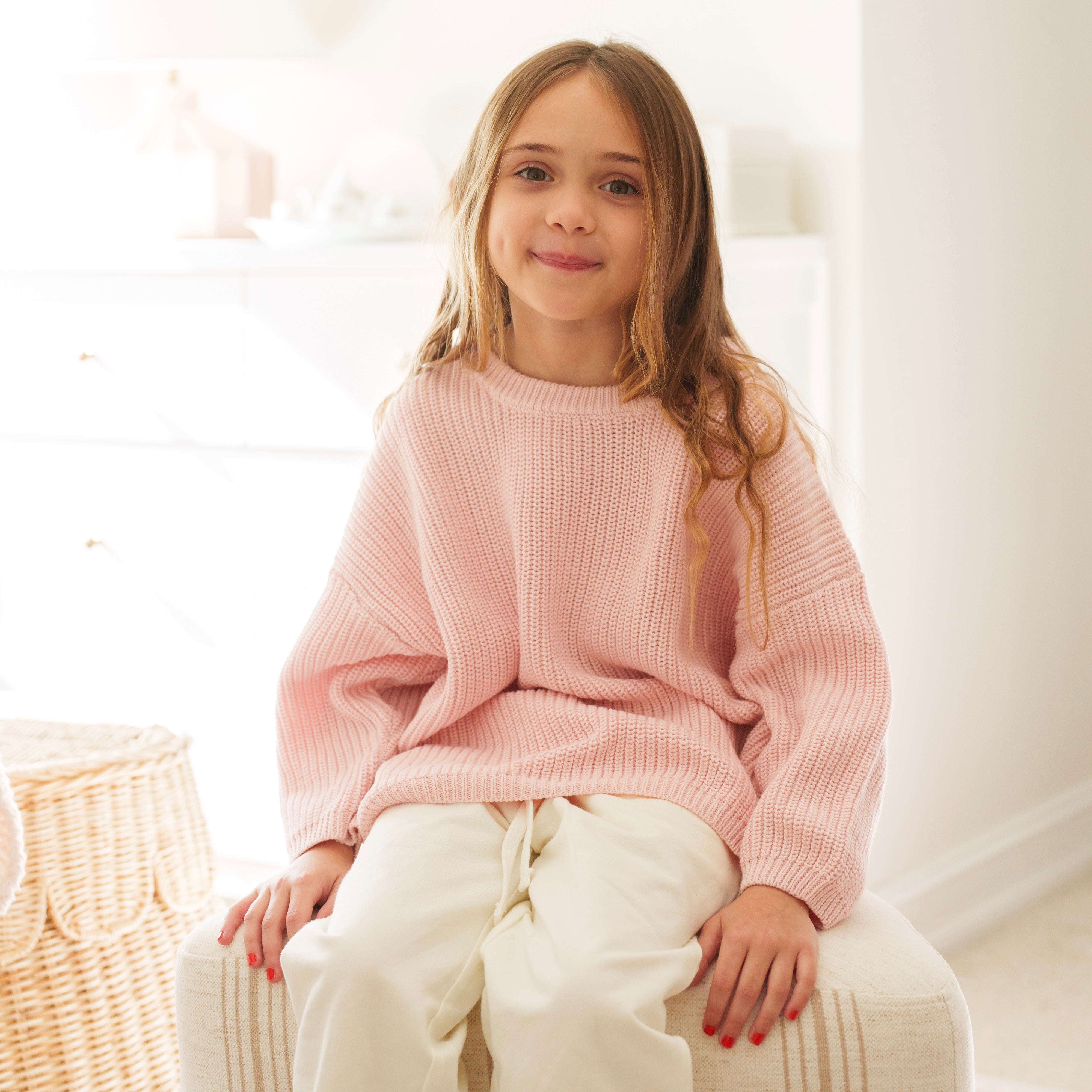 Girl wearing an Organic Chunky Knit Sweater in soft blush color, seated on a cozy ottoman.