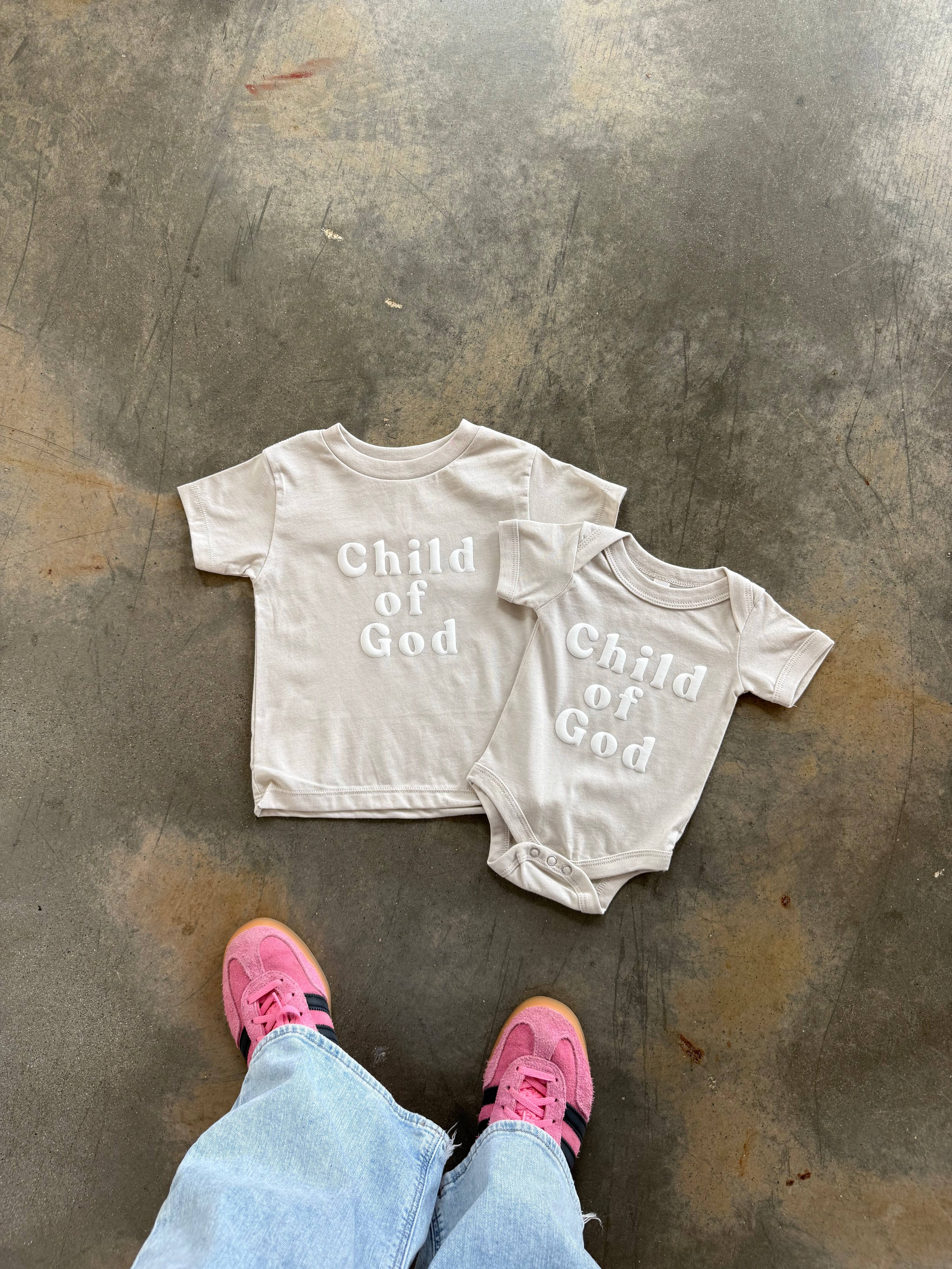 Child of God Onesie on a concrete floor with a person wearing pink shoes and blue jeans.