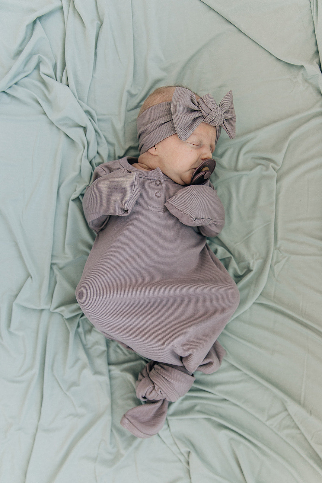 Baby wrapped in Bamboo Stretch Swaddle Sage, wearing a soft headband on a green background.