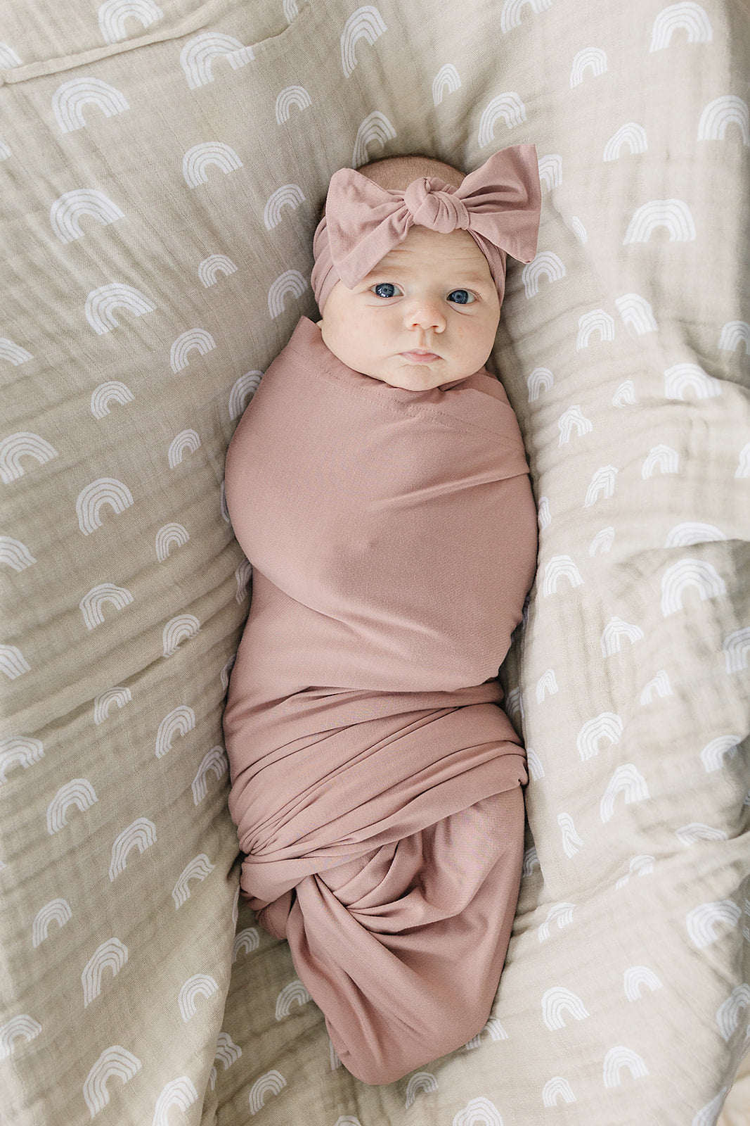 Bamboo Stretch Swaddle Dusty Rose wrapping a baby with a bow headband on a cozy blanket.