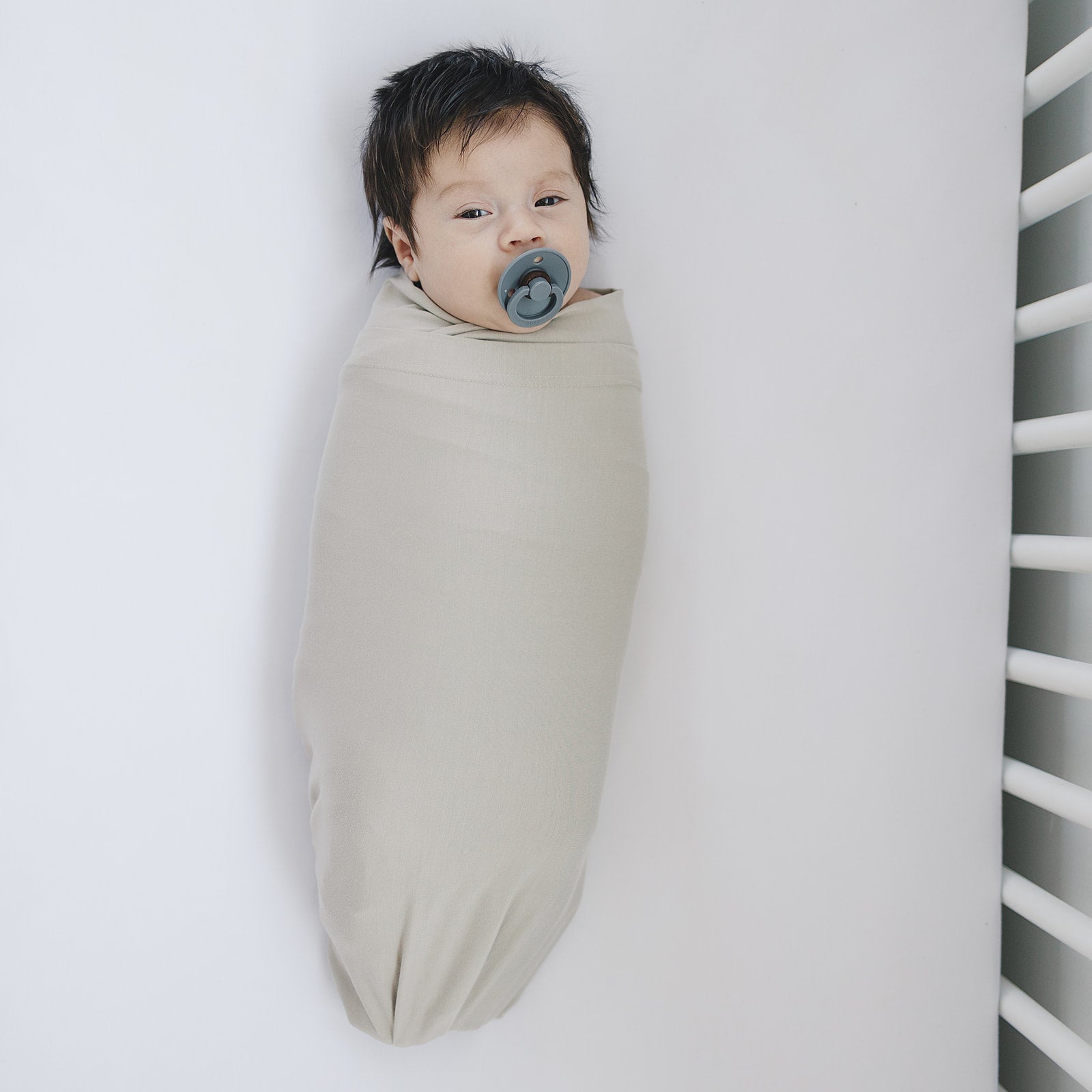 Bamboo Stretch Swaddle Oatmeal wrapped around a peaceful baby with a pacifier in a minimalist crib setting.
