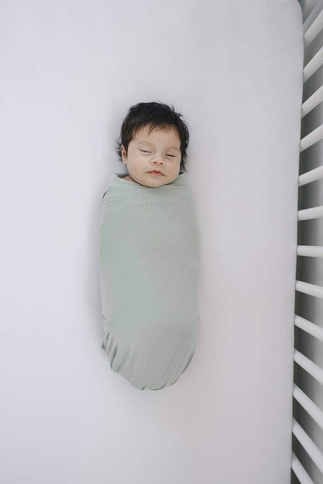Baby wrapped in Bamboo Stretch Swaddle Sage on a crib mattress, showcasing soothing sage color and cozy design.
