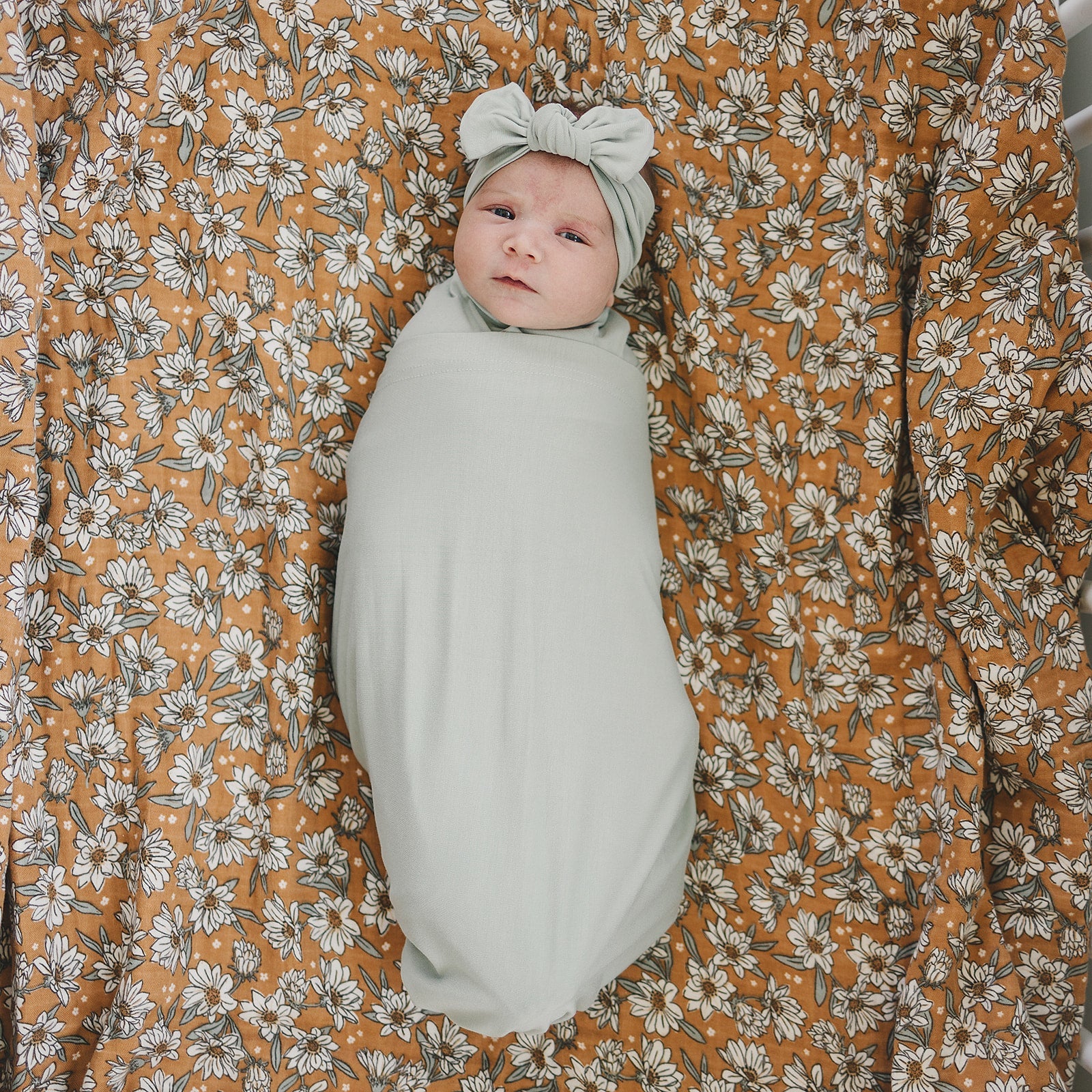 Baby wrapped in Bamboo Stretch Swaddle Sage on a floral patterned blanket.