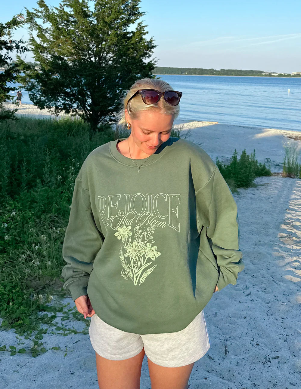 Woman wearing a forest-green Rejoice unisex crewneck sweatshirt by the beach, featuring floral design and relaxed fit.