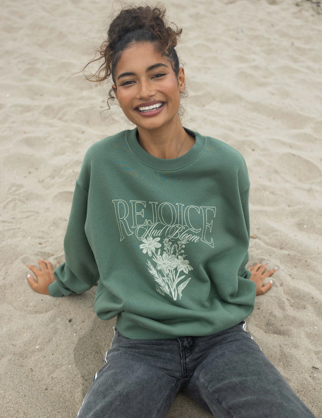 Woman smiling in a forest-green Rejoice crewneck sweatshirt sitting on the beach, showcasing faith-inspired design.