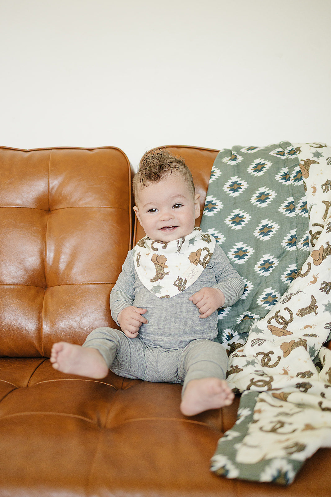 Baby wearing Outlaw Cowboy muslin bib, sitting on a brown couch with stylish patterned blanket.