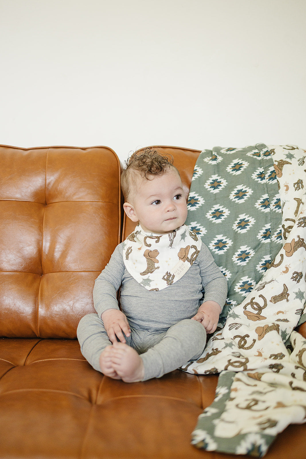 Baby wearing Outlaw Cowboy Classic Muslin Bib by Mebie Baby while sitting on a brown leather couch.