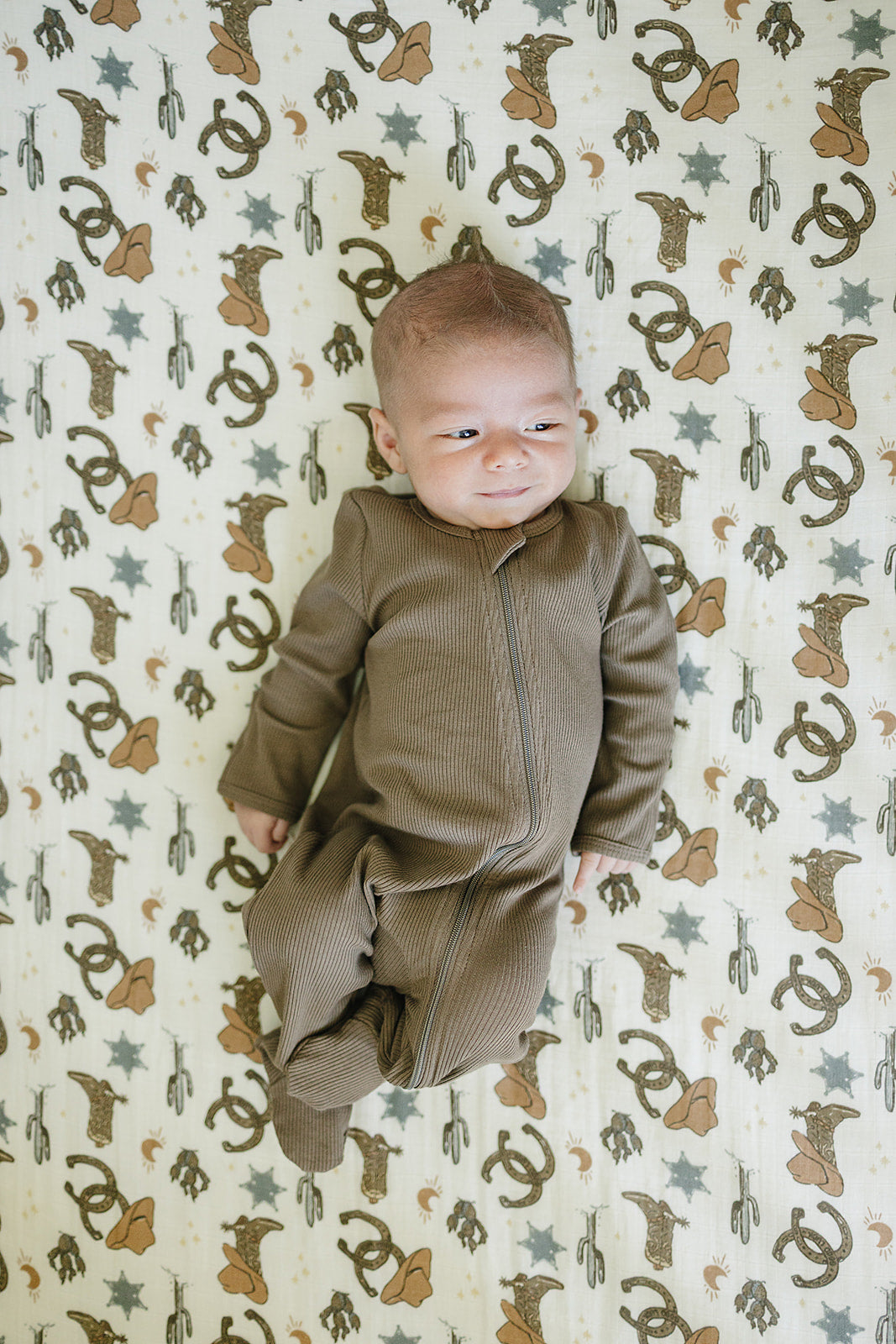 Muslin crib sheet featuring playful outlaw cowboy design with a baby in a brown zip-up onesie.