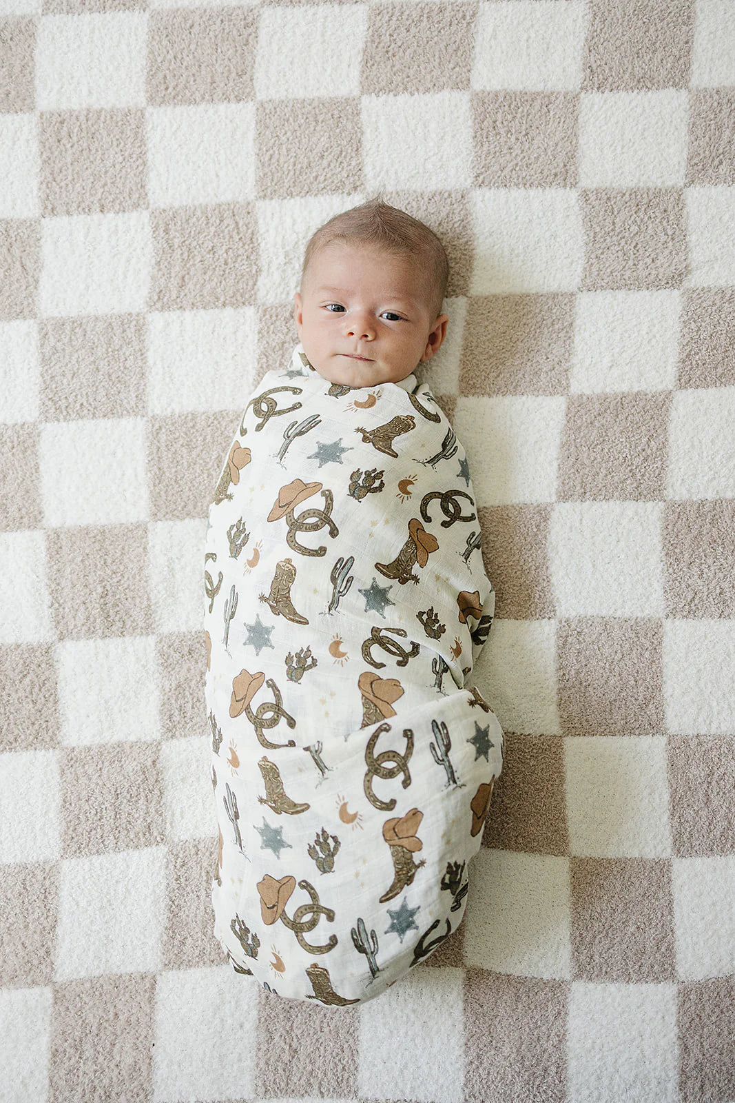 Baby swaddled in outlaw cowboy muslin swaddle blanket on a patterned rug, showcasing playful Western design.