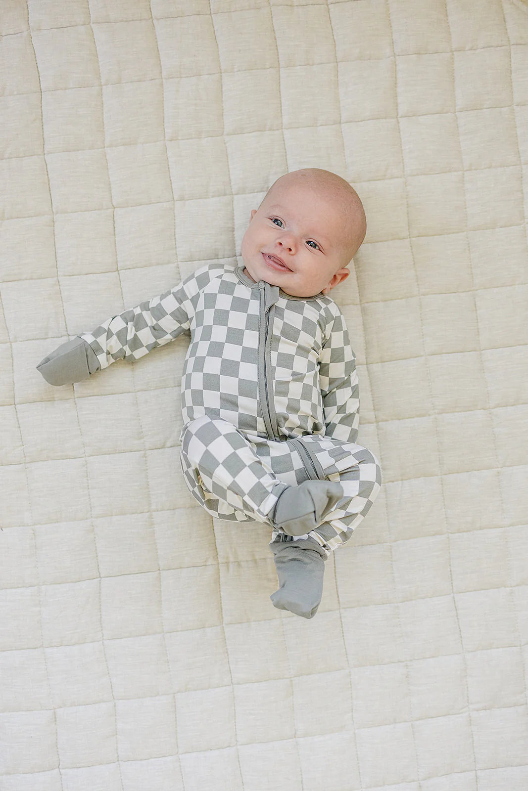 Baby in light green checkered bamboo zipper pajama on a quilted blanket, smiling cheerfully.