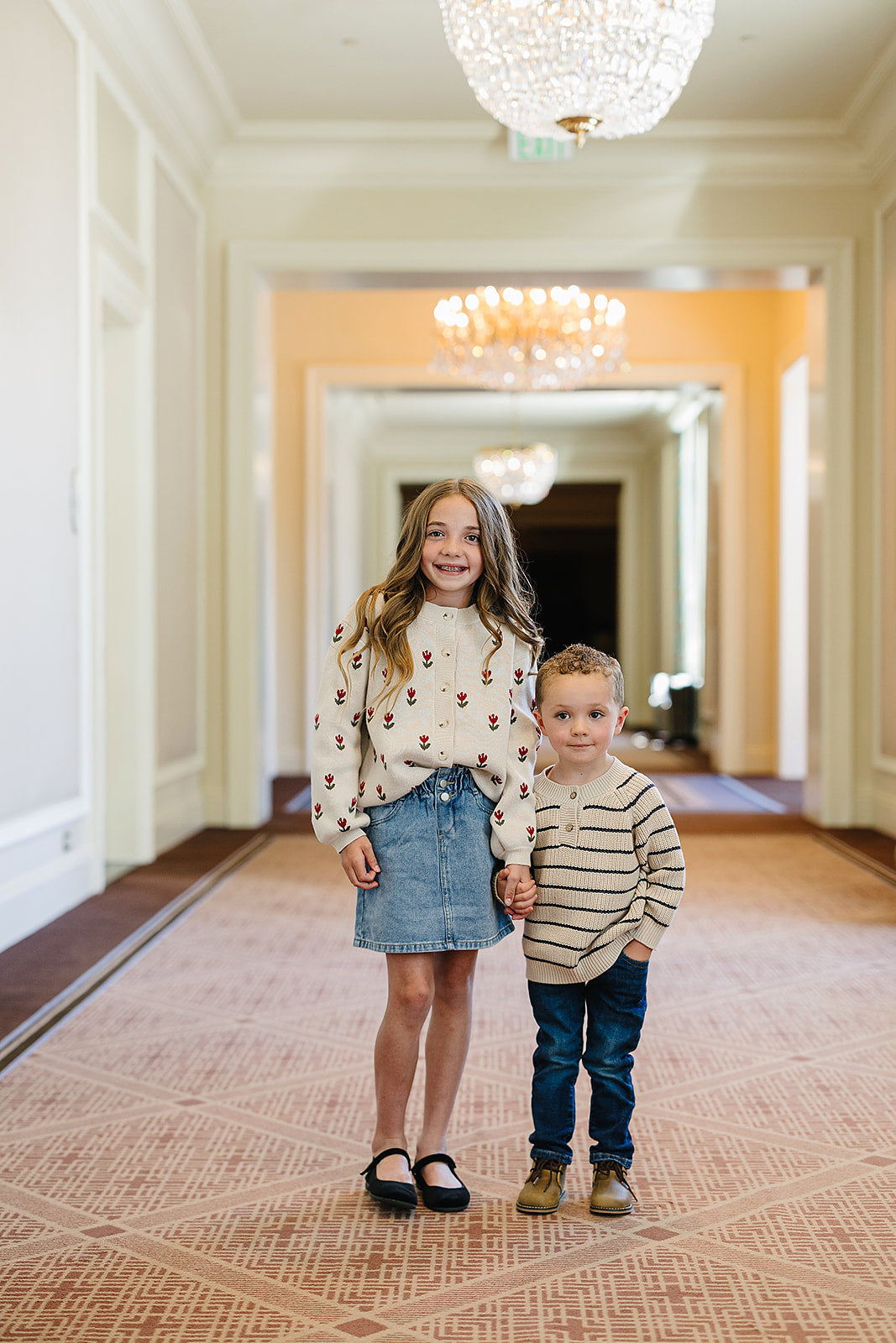 Two kids standing in a hallway, the girl wearing a classic knitted baby sweater and the boy in a striped infant top.